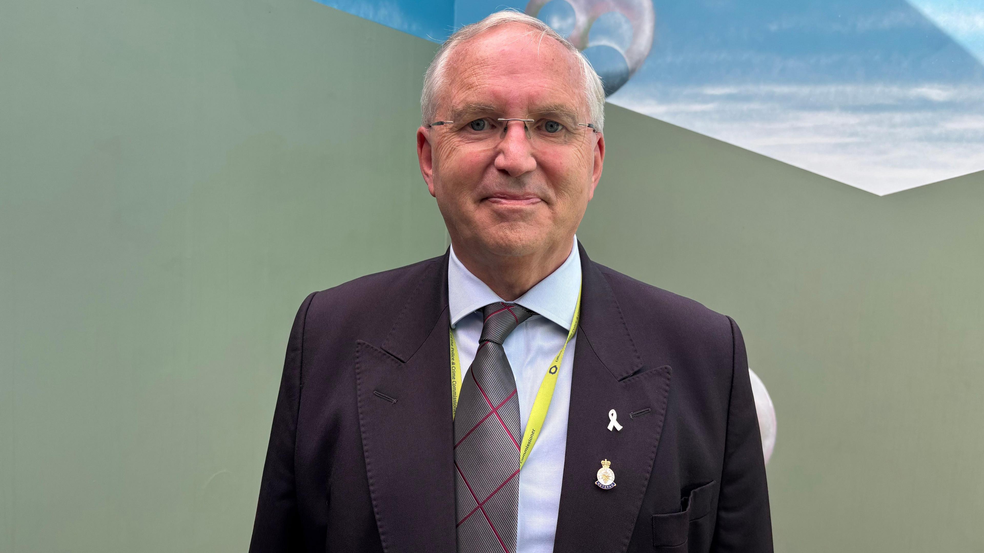 Police and Crime Commissioner Chris Nelson is dressed in a dark suit and stands in front of the mural which features an olive green base and a daylight skyline above him. 