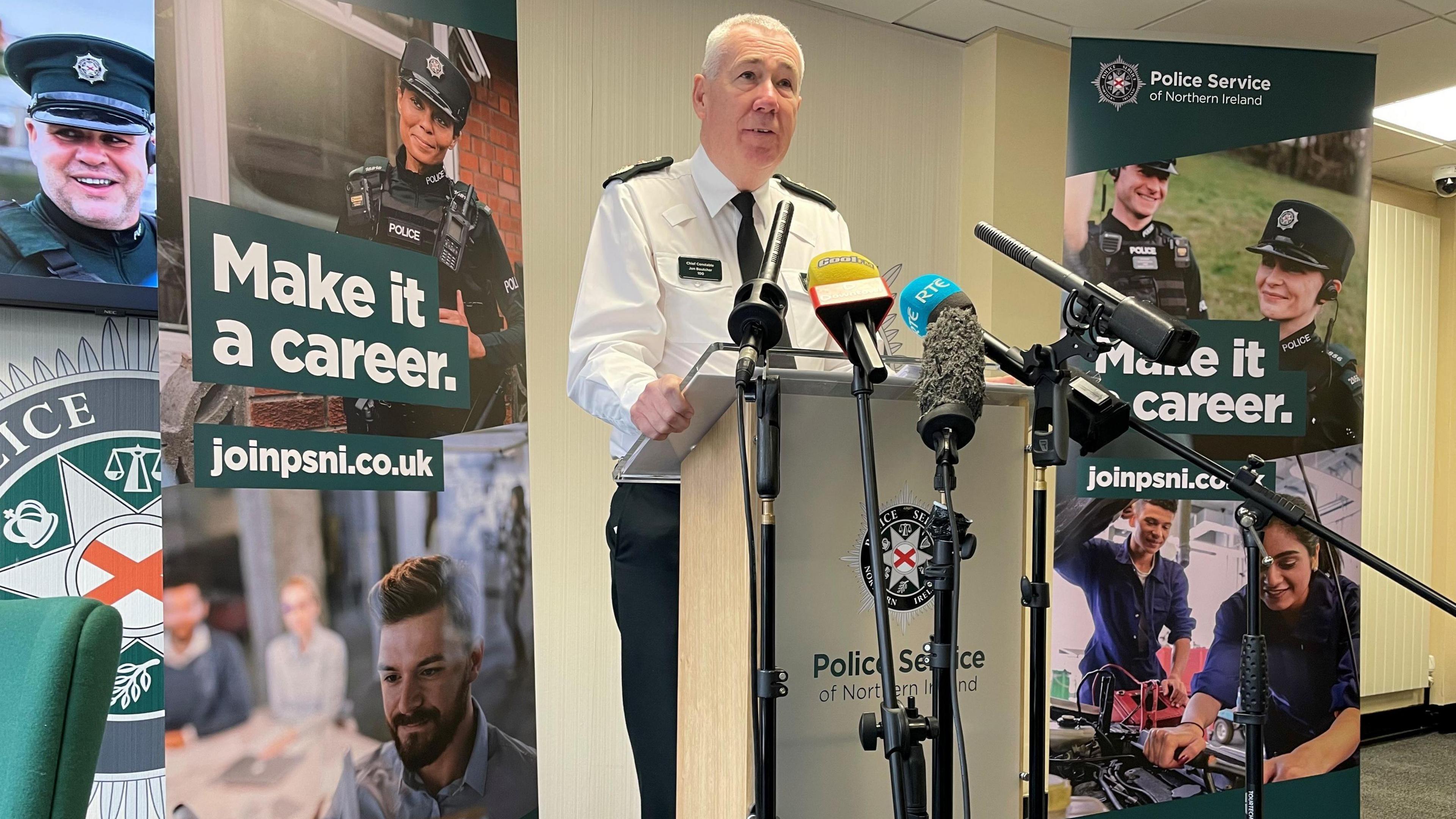 A grey haired man in a white shirt, green trousers and black tie, stands at a wooden plinth speaking into six microphones. On the front of the plinth is a circular badge with grey, green and black colours. Either side of him, behind are two seven foot posters with the words 'make it a career' joinpsni.co.uk. The poster shows recruits helping members of the public. 