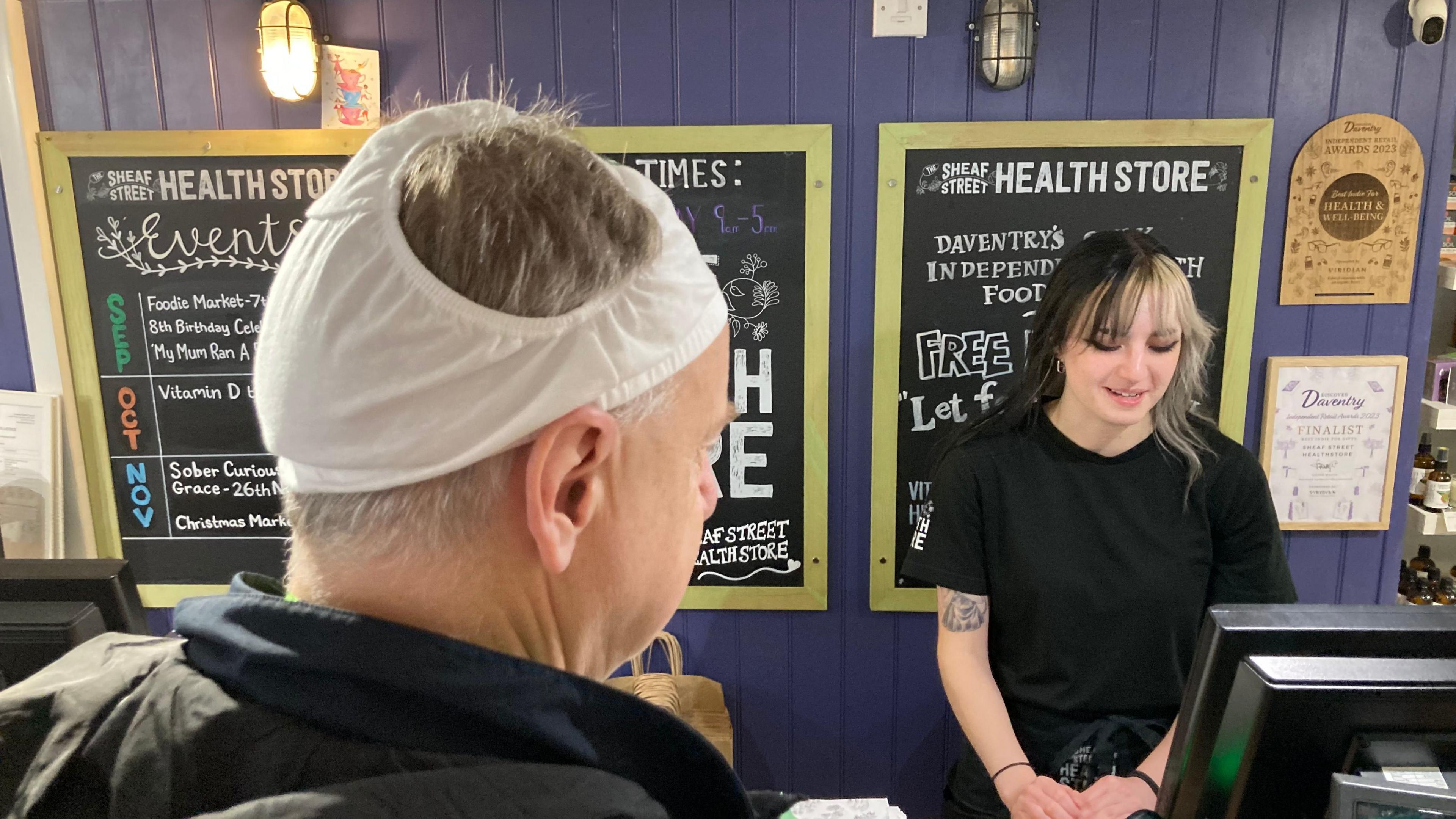 Mark Robson, with short grey hair and white pants on his head, looks at Oliver Hall, with long hair which is dark on one side and light on the other. Oliver is wearing a black T-shirt and is standing behind a till.  She is in front of blackboards showing some of the items on sale in the shop.