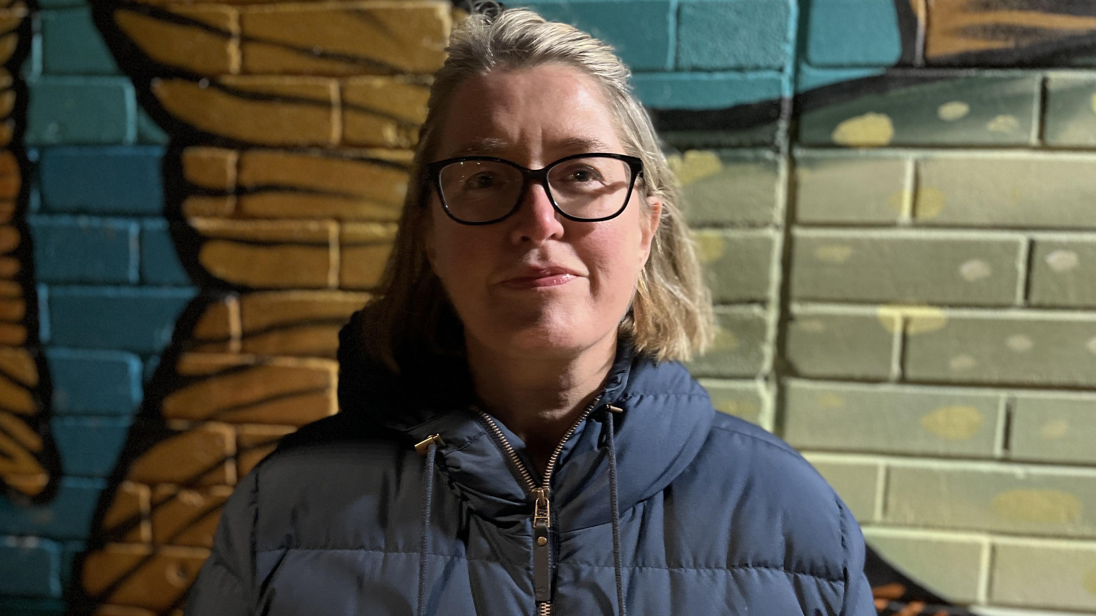 Annie Hubbard, Encircles trustee and volunteer, standing against a brick wall wearing large black rimmed glasses and bobbed blonde hair.