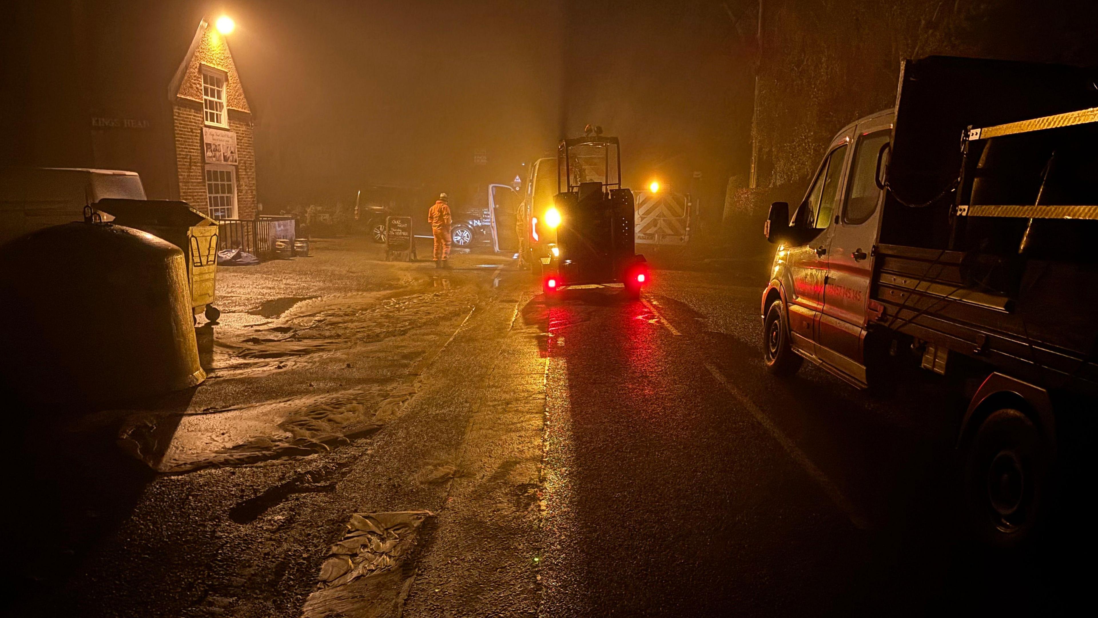 A street lit by streetlamps is flooded. It is dark but the lights of several maintenance trucks can be seen. 