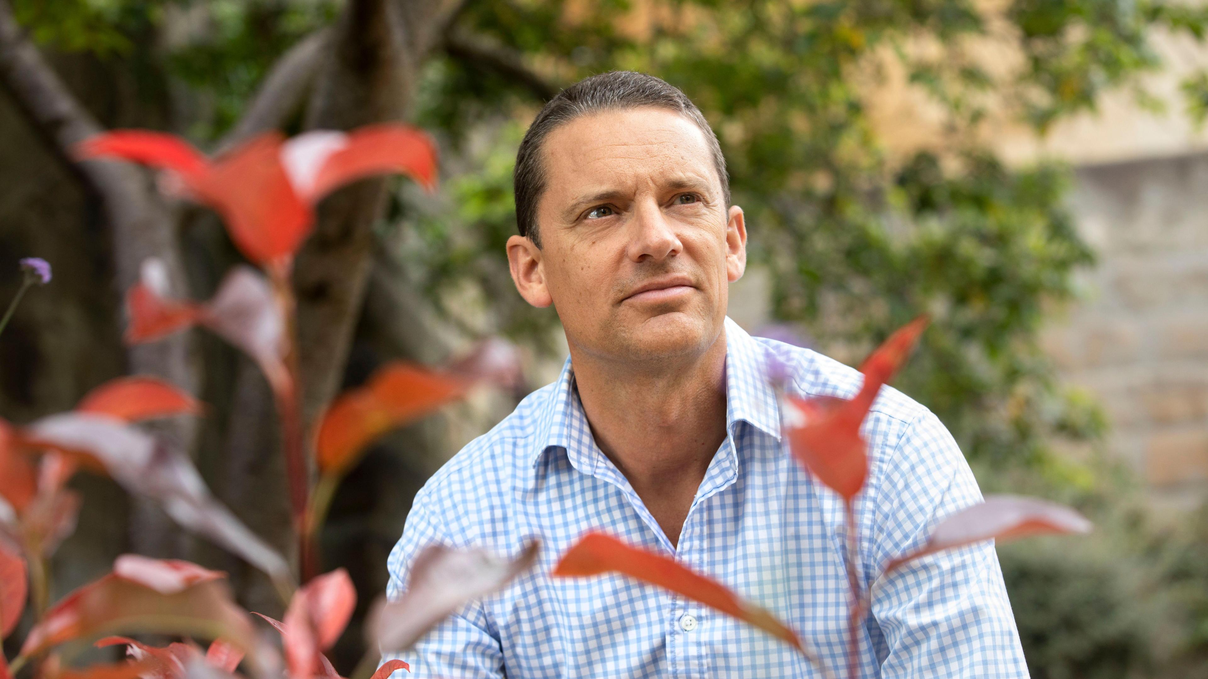 Bevis Watts in a blue and white checked shirt sitting amongst trees and plants