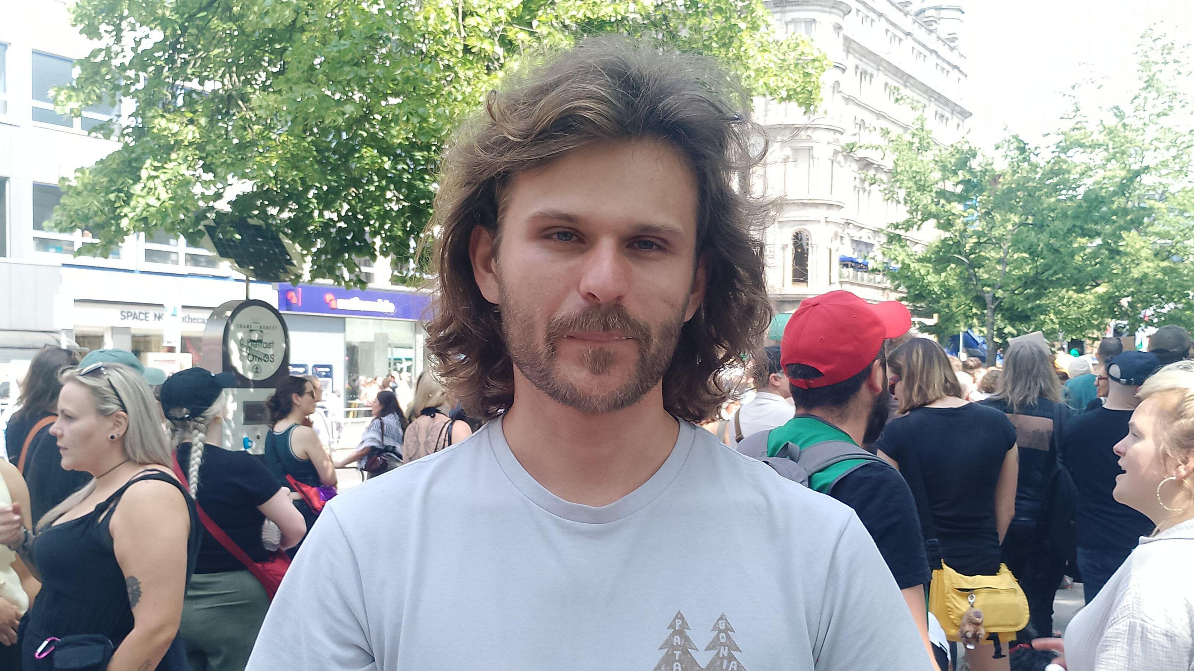 A man, with shoulder length brown hair and brown stubble, looking at the camera. He is wearing a light blue t-shirt. There is a crowd of people behind him.