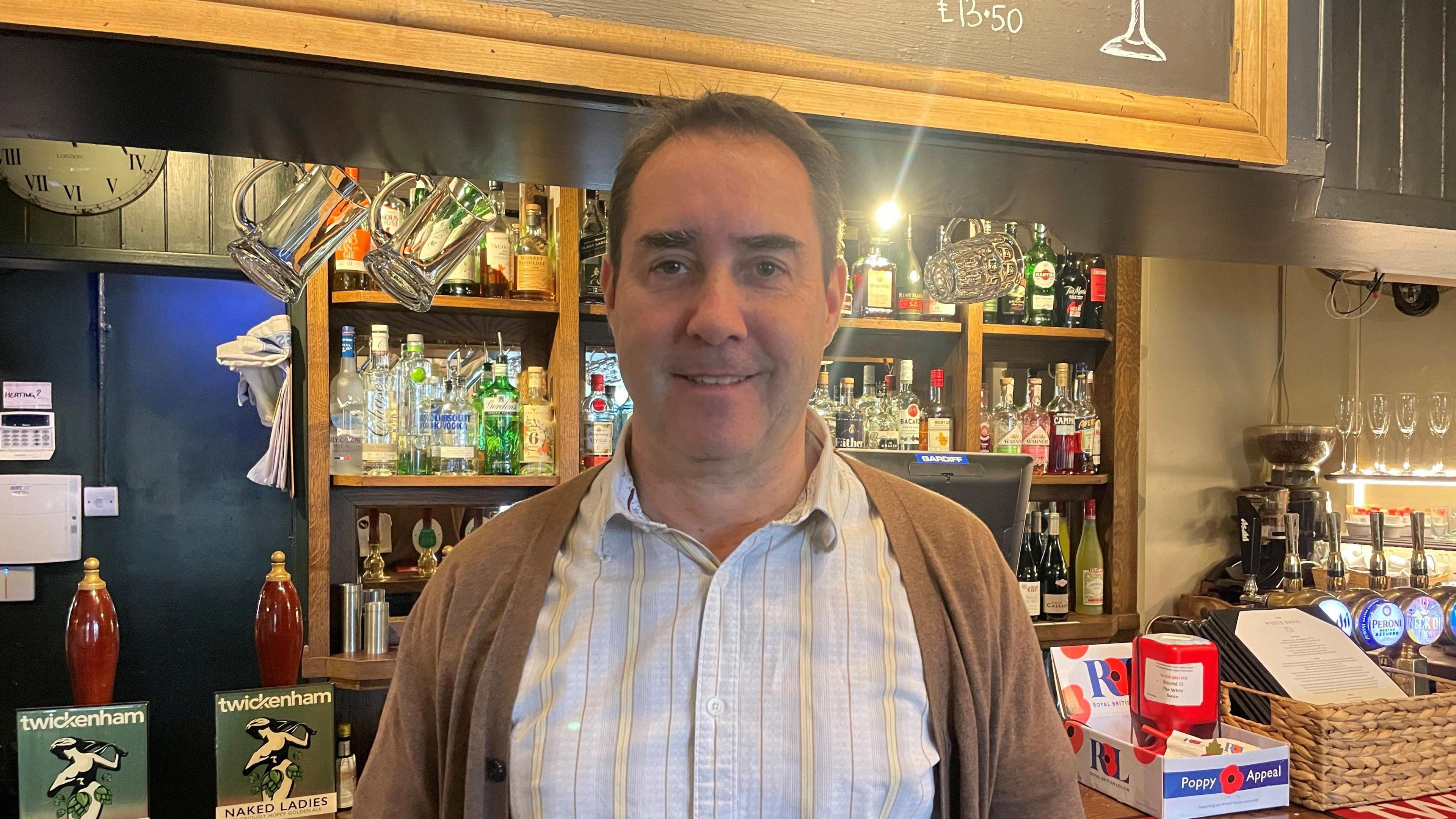 'The Poisoned Swan', pub manager Kevin O'Callaghan wearing a blue and yellow striped shirt and brown cardigan. he is stood in front of the bar, with glassware and bottles behind him