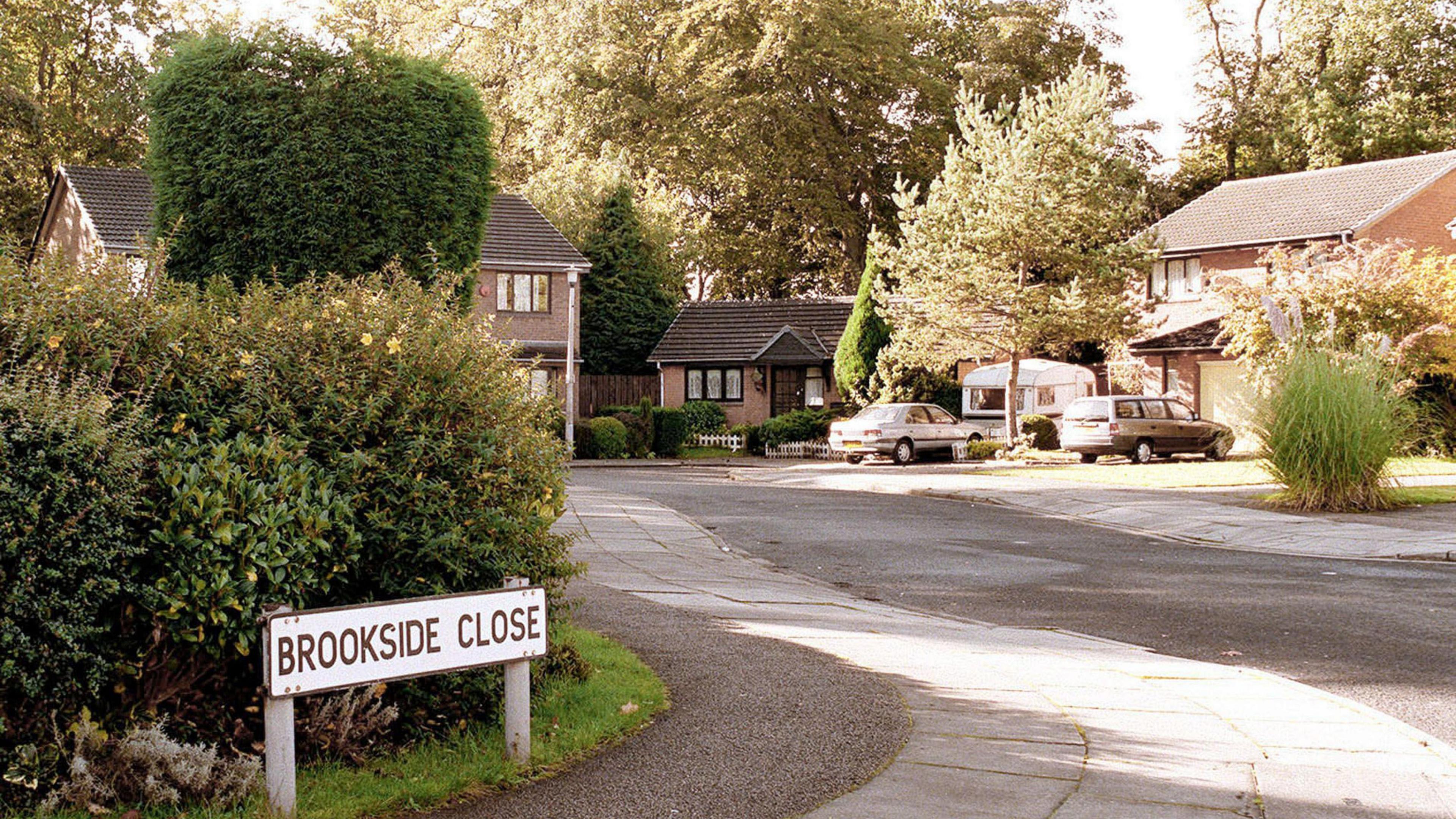 A picture of Brookside Close, captured in the 1990s. To the left is a street sign which reads: Brookside Close with bushes behind it. The road curves round to the left with a number of houses and one bungalow in view.