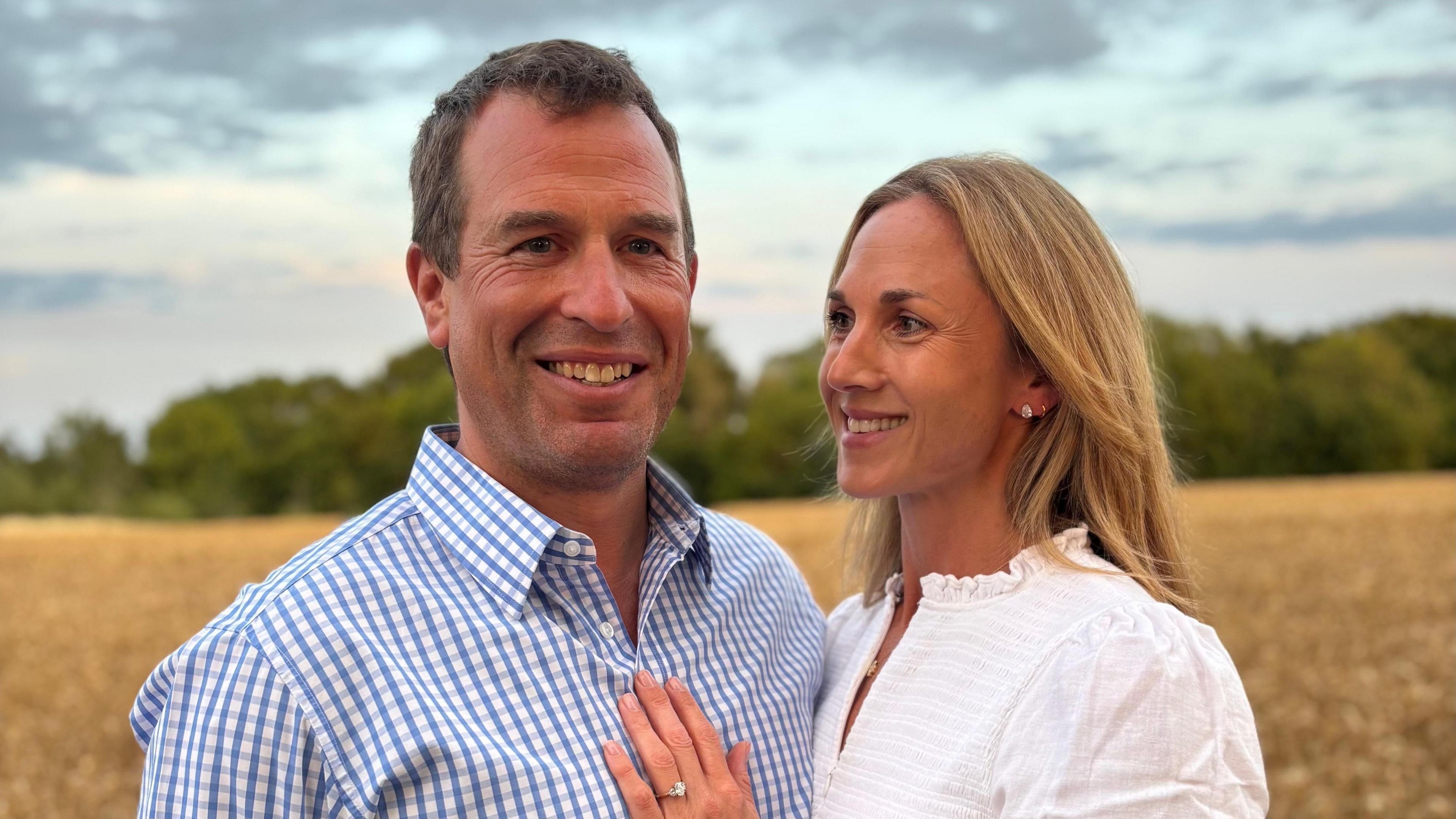 Peter Phillips wheres a checkered shirt and Harriet Sperling in a white blouse puts her hand with engagement ring on her fiance's chest. They stand in a field.