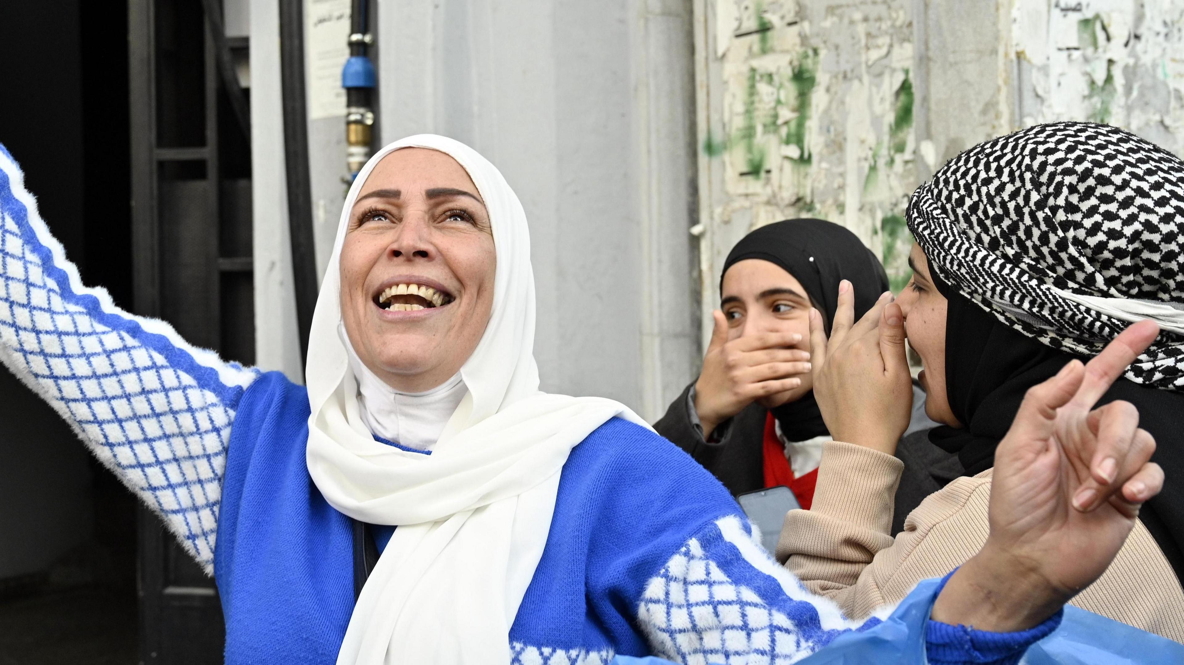 A smiling woman in a blue and white dress looks up to the sky. Behind her two other women cover their smiles with their hands.