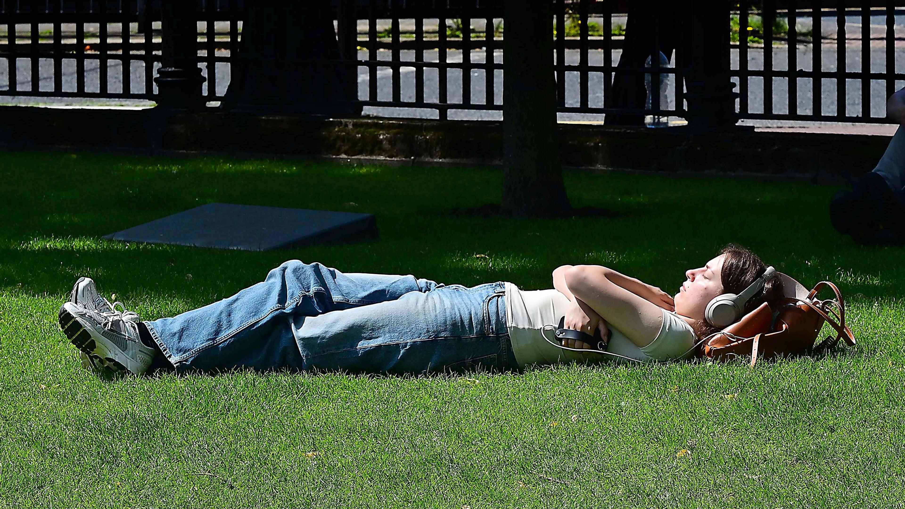 A woman wearing headphones lying on grass with her arms crossed and eyes closed. She has dark hair, wearing a white t-shirt, jeans and trainers.