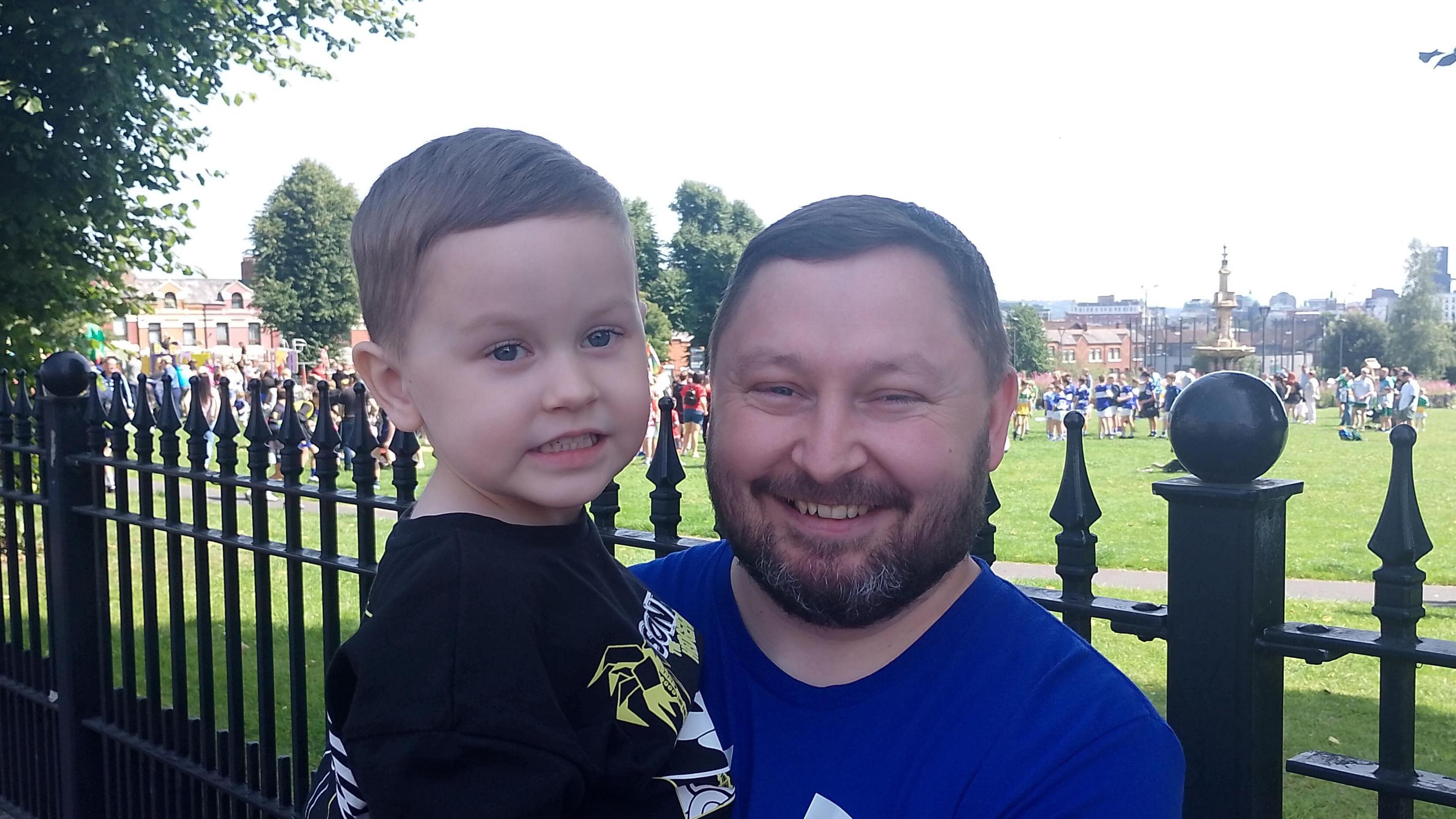 Kevin, wearing a blue t-shirt, smiles at the camera as he holds a young child.