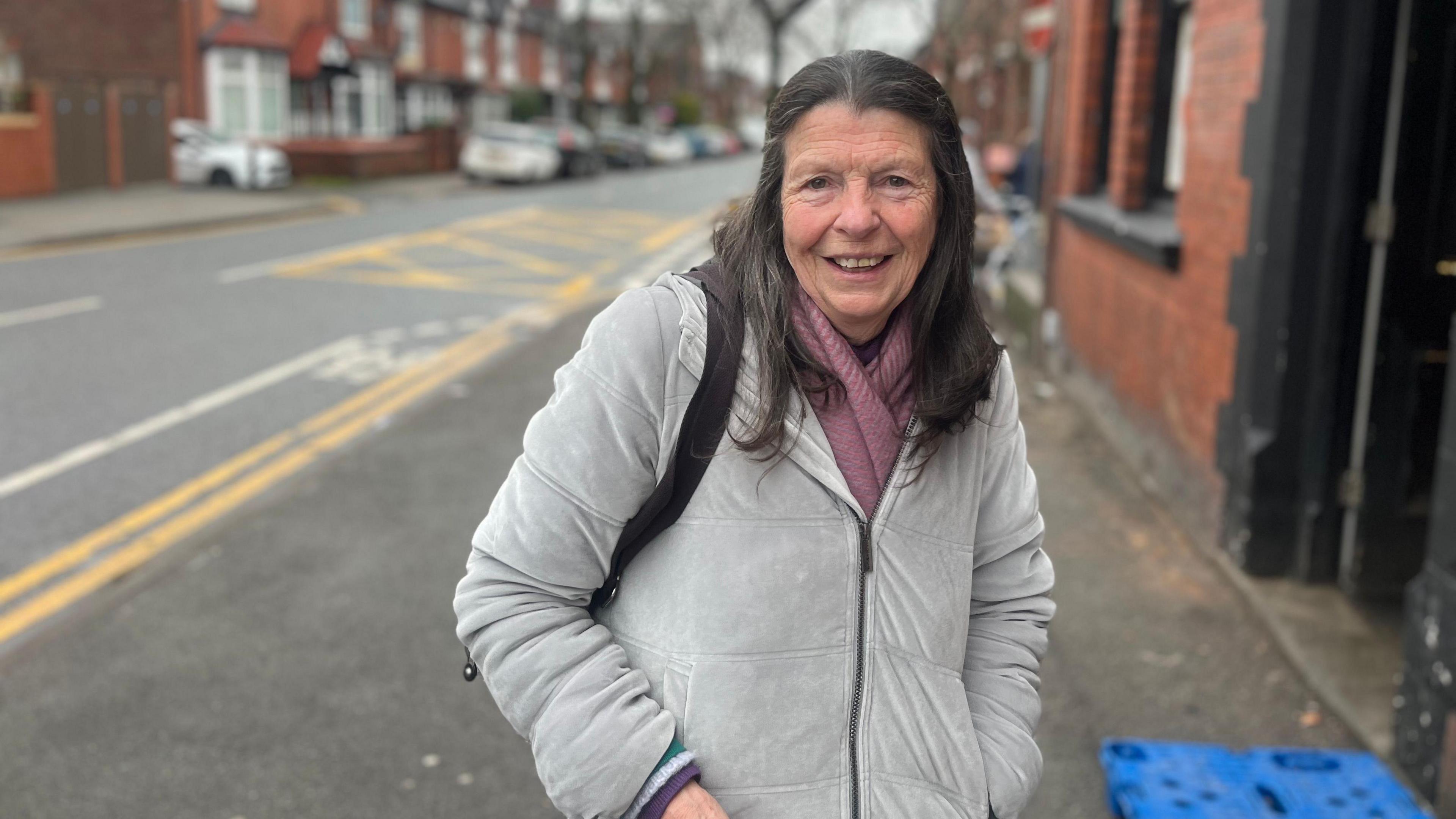 Joan Good is smiling as she stands outside the shop. She is wearing a grey jacket and a pink scarf and she has long brown hair.