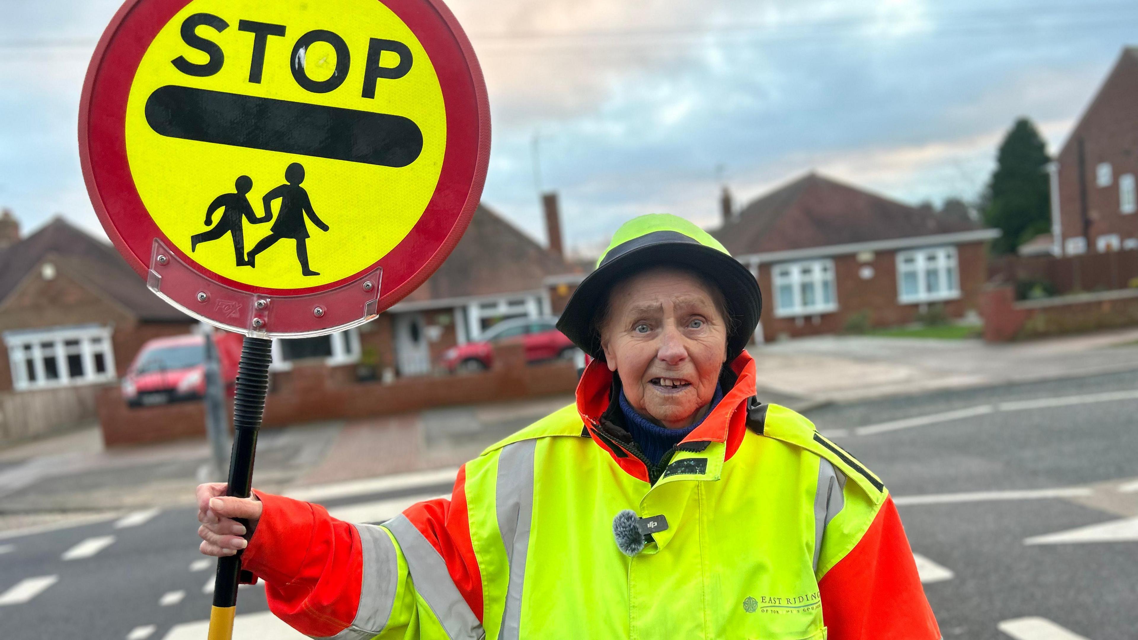 An elderly lady wearing a high-vis uniform holding her lollipop next to the zebra crossing she has worked on for 40 years