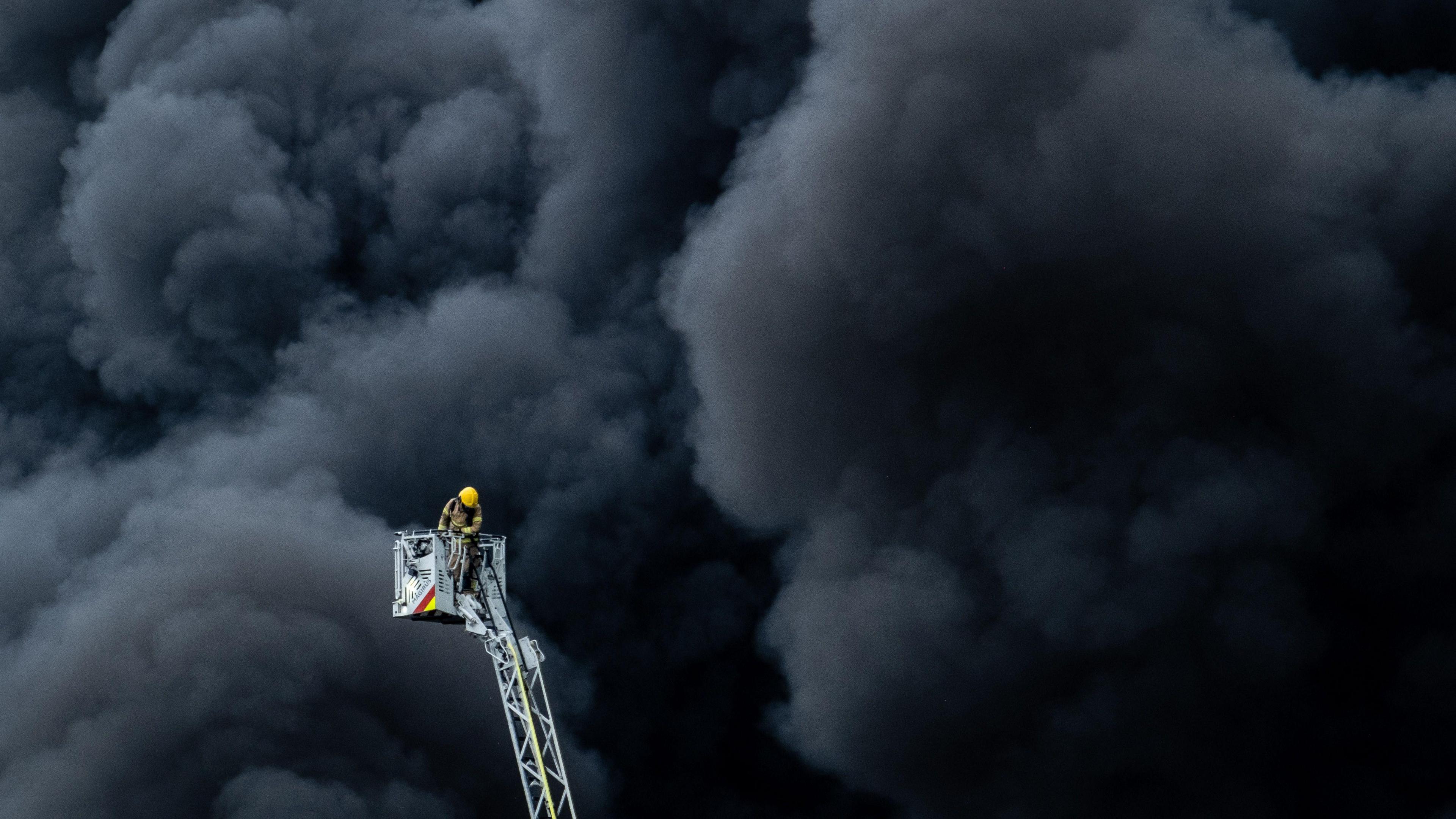 A fire fighter on a ladder stands in front of a blanket of thick black smoke