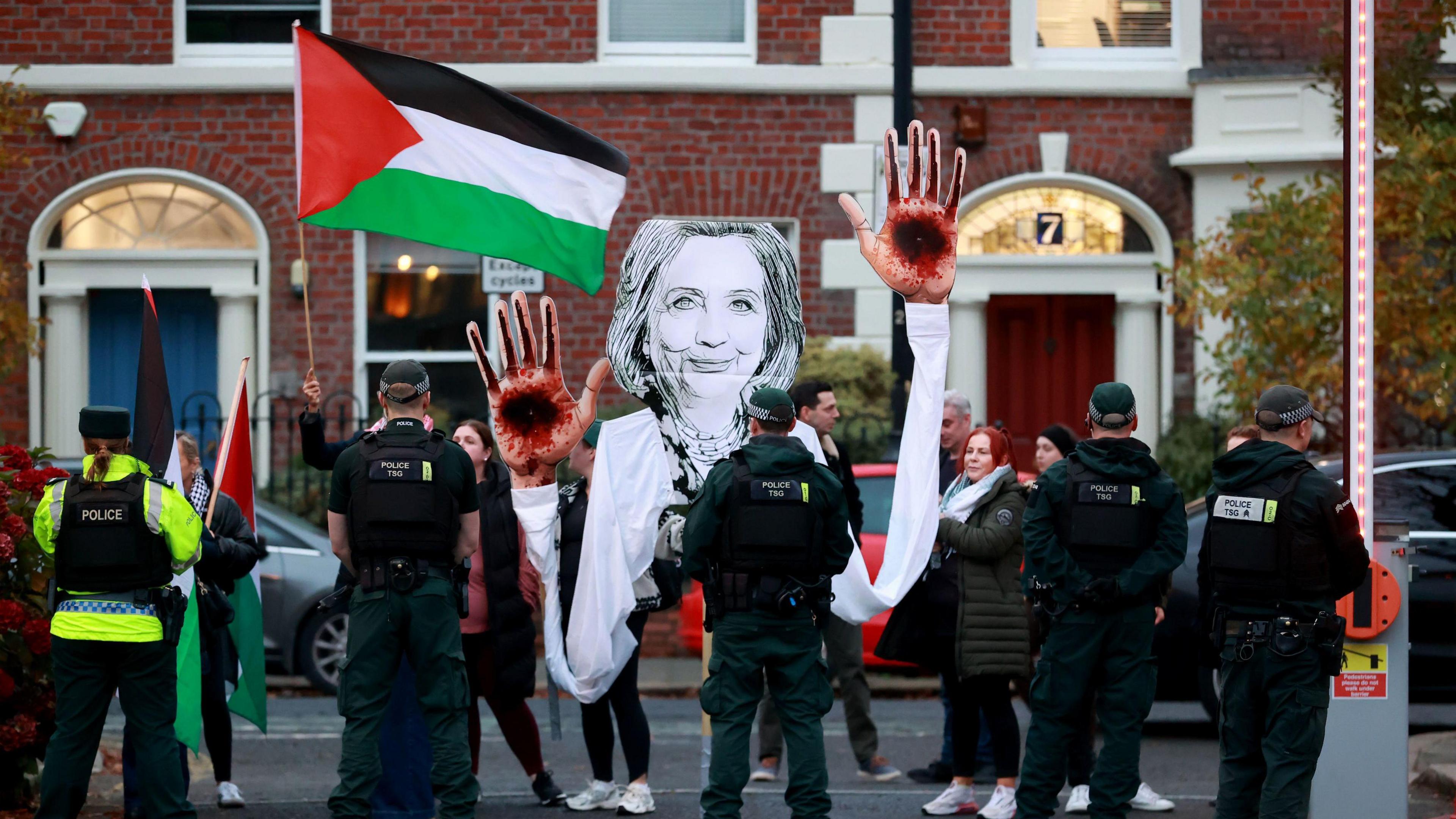 Protestors are pictured standing behind police holding a Palestinian flag and a large black and white cut out of Hilary Clinton with blood on her hands.