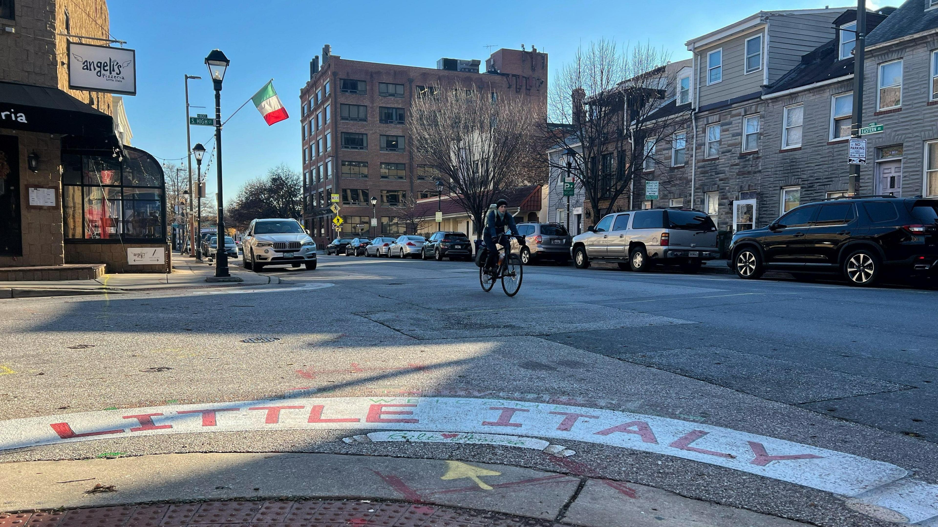 The foreground is a street with Little Italy painted on it and the background shows older brick buildings and a man bicycling down a street.