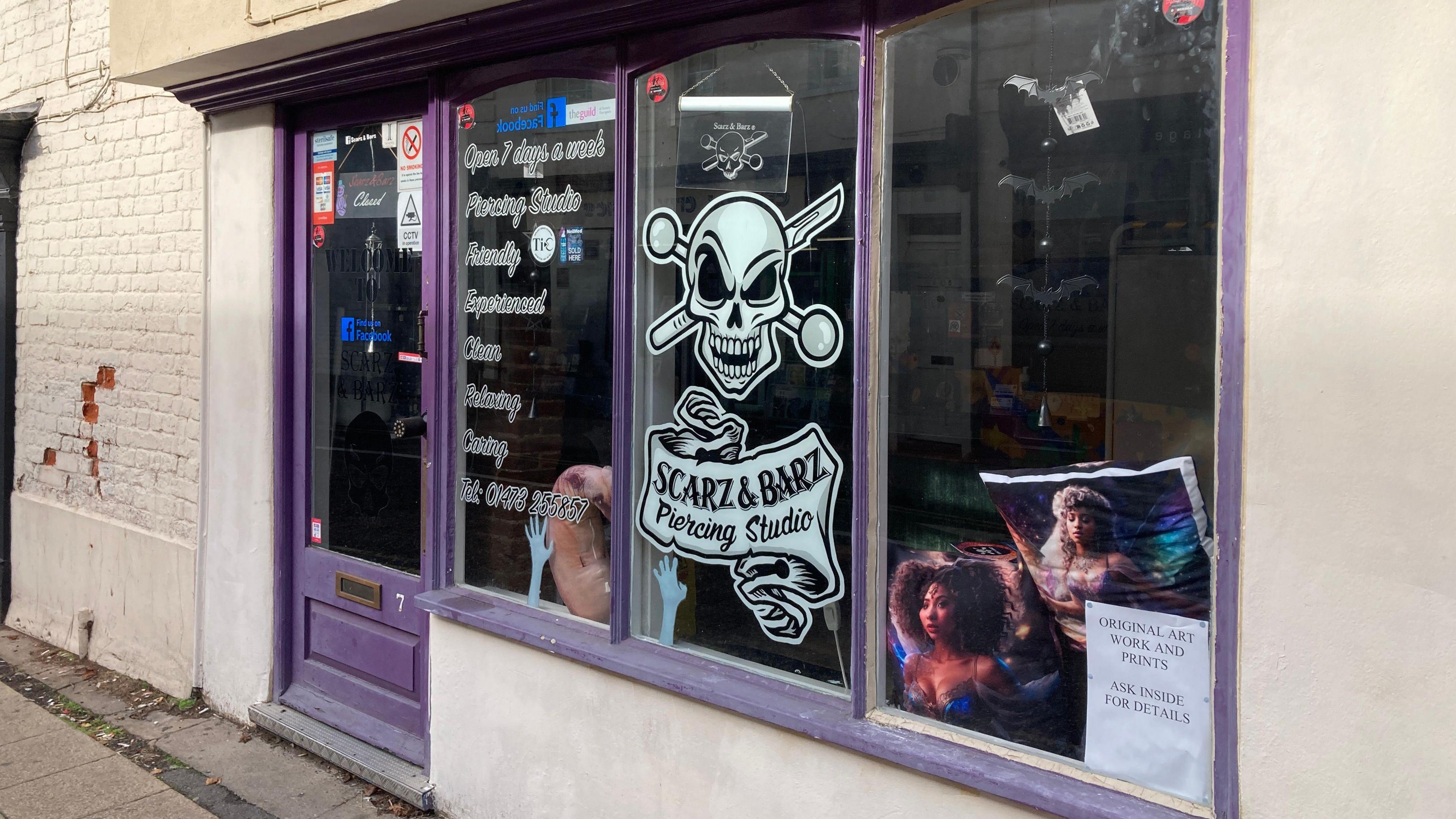 The Scarz&Barz piercing studio has purple framed windows and a front door. Its skull log is on the central window in white. The surrounding brickwork/plaster is painted white.

