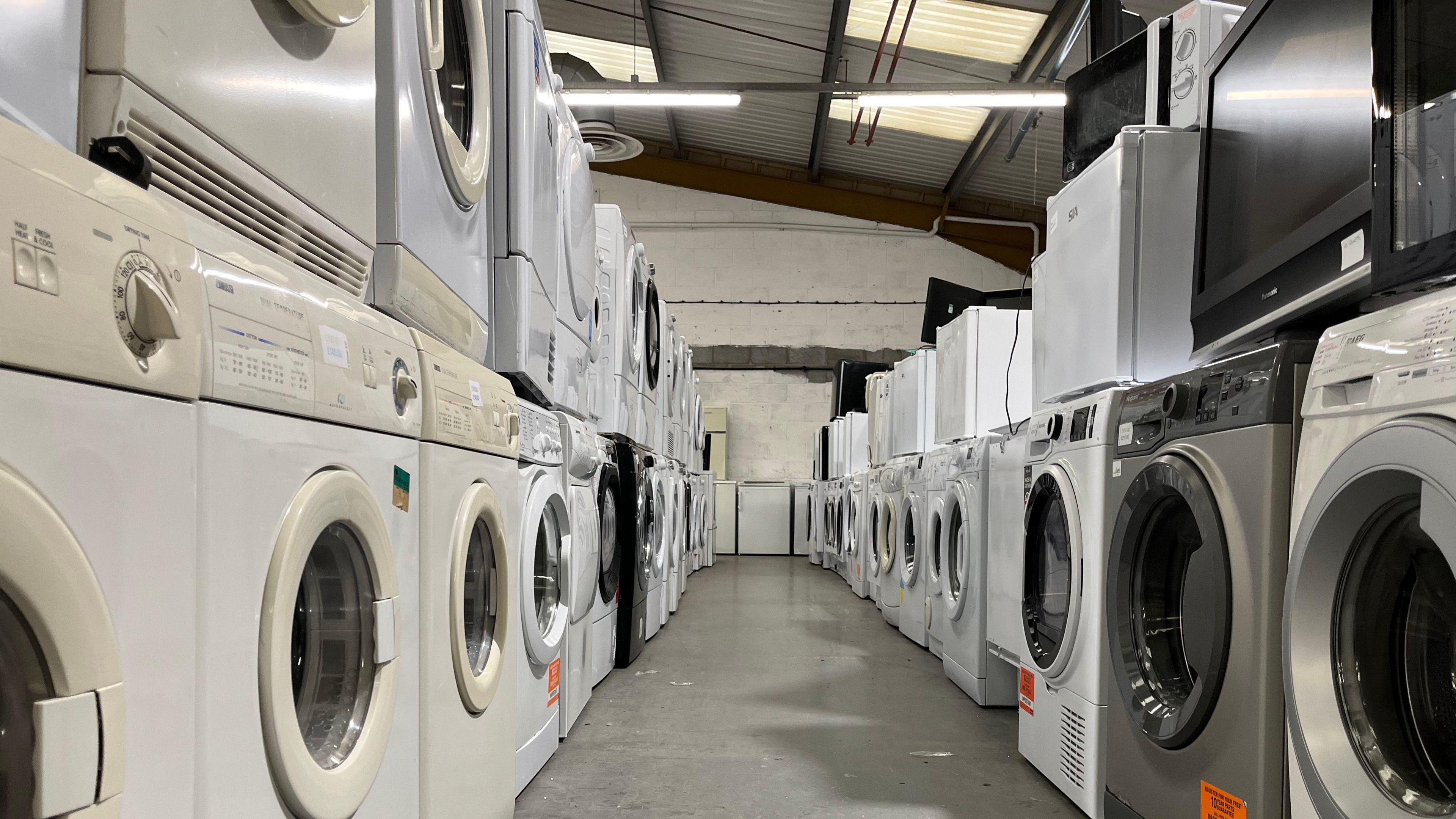 Looking down two rows of washing machines stacked in a warehouse. They are all different as they are second-hand. 