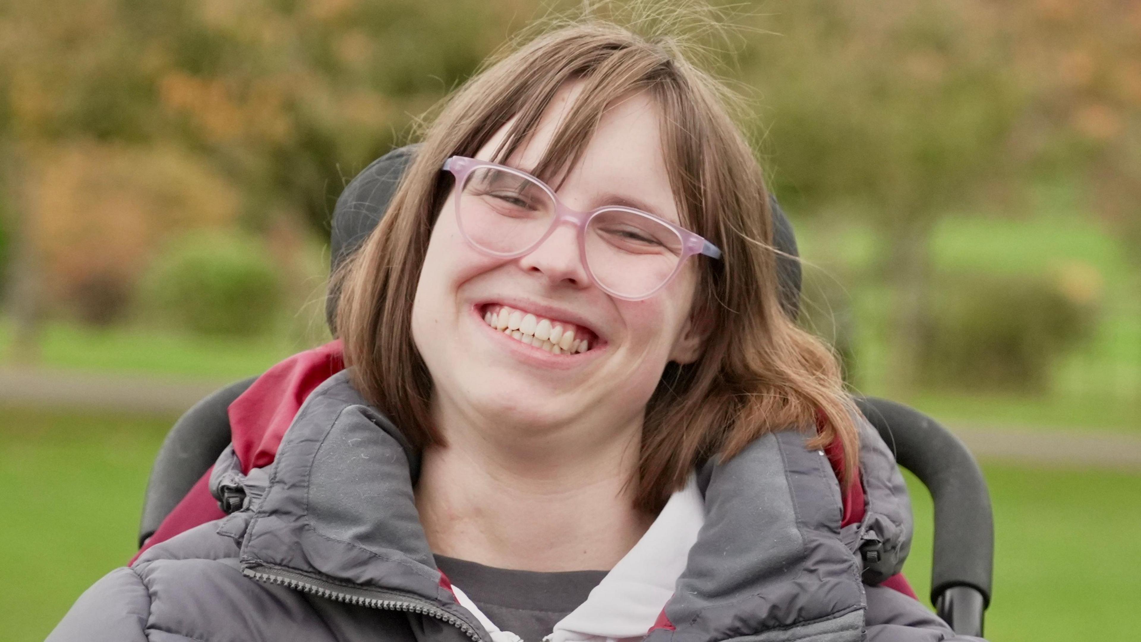 Kate is a young women in a wheelchair. She has brown shoulder length hair and wears glasses. She has on a grey puffa jacket and is smiling broadly.
