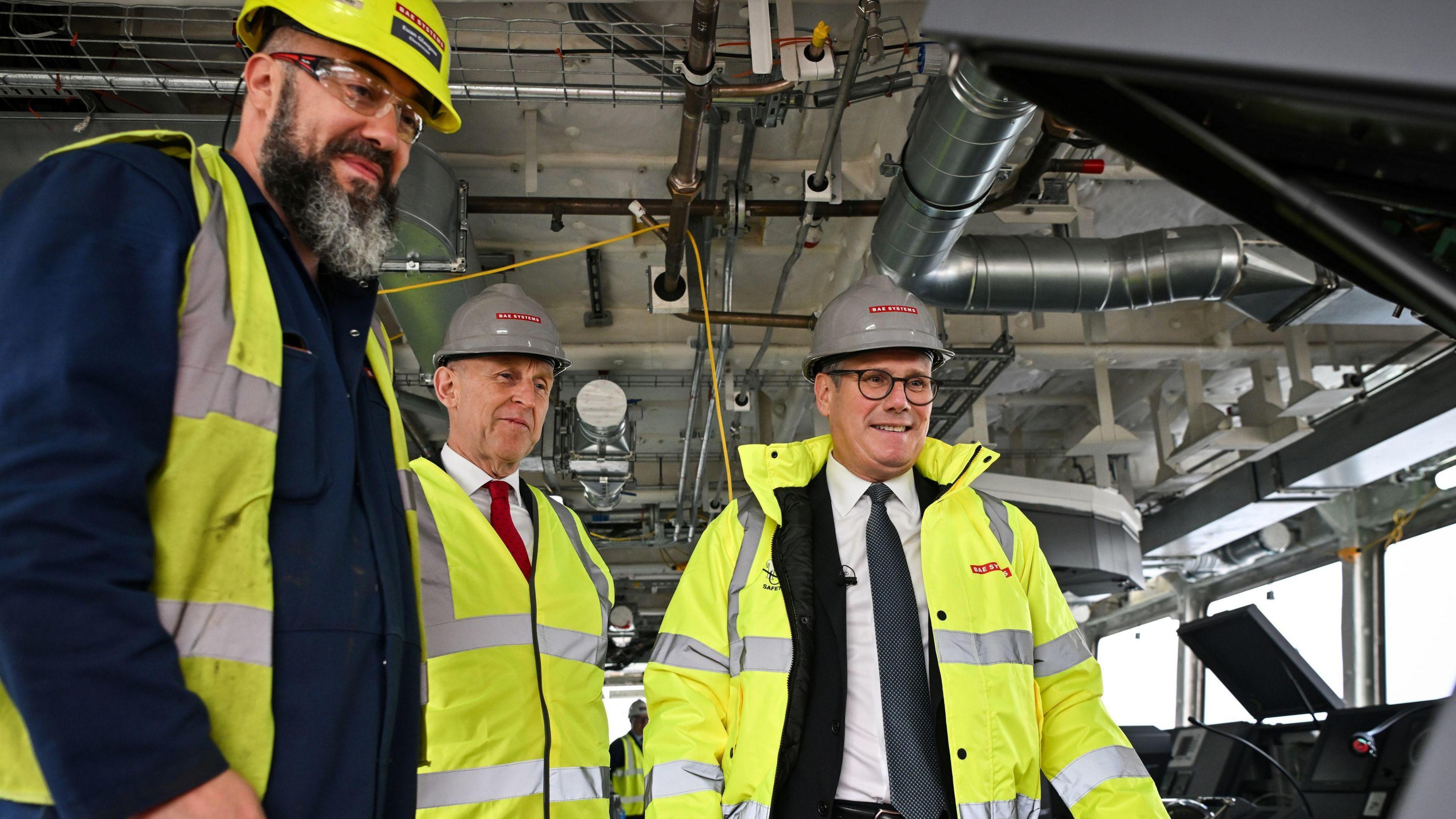 Defence Secretary John Healey (centre) and Prime Minister Sir Keir Starmer (right) during a visit to BAE Systems in Govan, Glasgow, to launch the Strategic Defence Review wearing high-visibility jackets and hard hats stand in an industrial setting, appearing to discuss or inspect machinery. The background features pipes, cables, and industrial equipment.