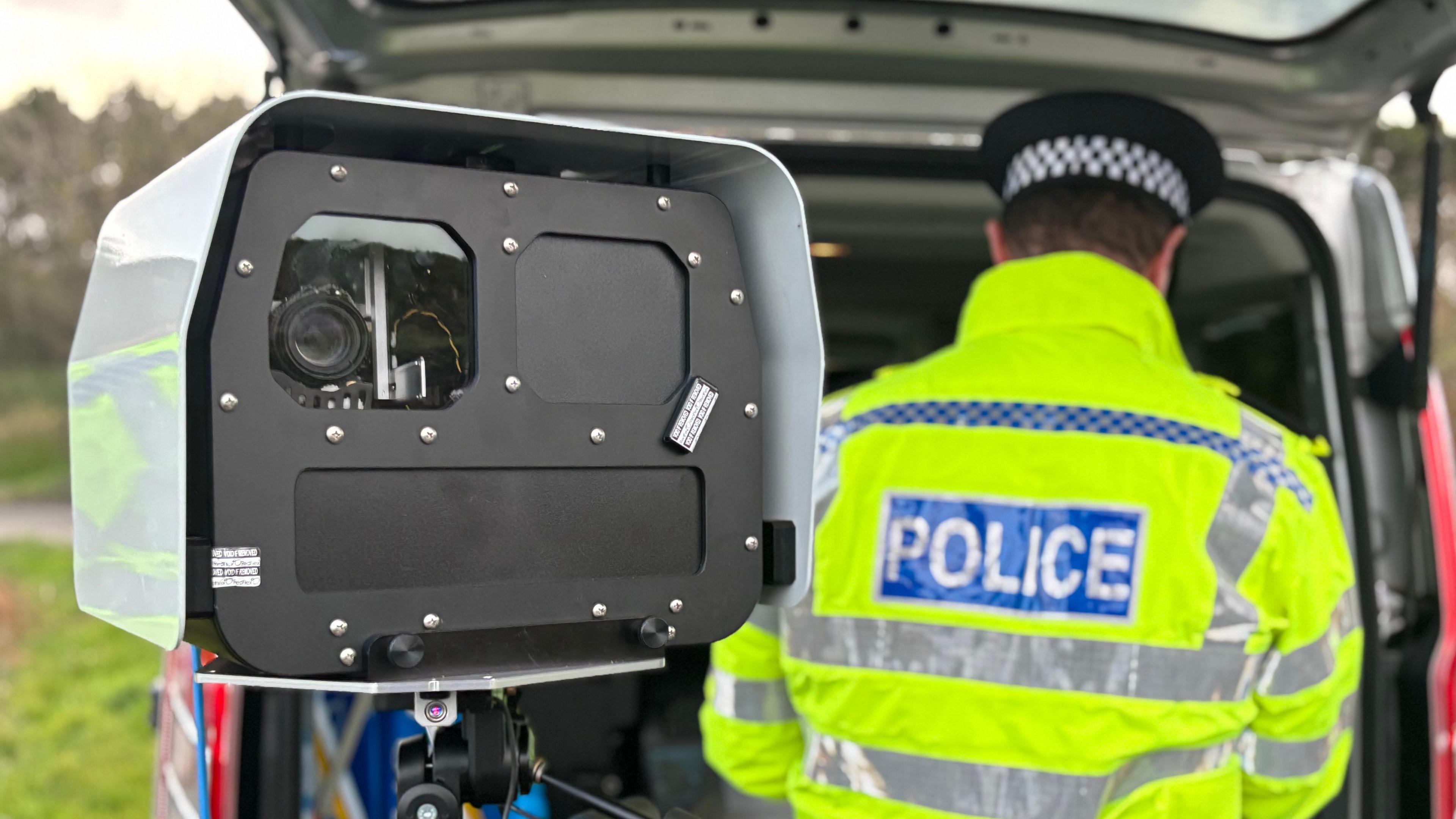A large rectangular speed camera on a tripod is visible. Behind it, a police officer in a high visibility jacket is working in a police van. 