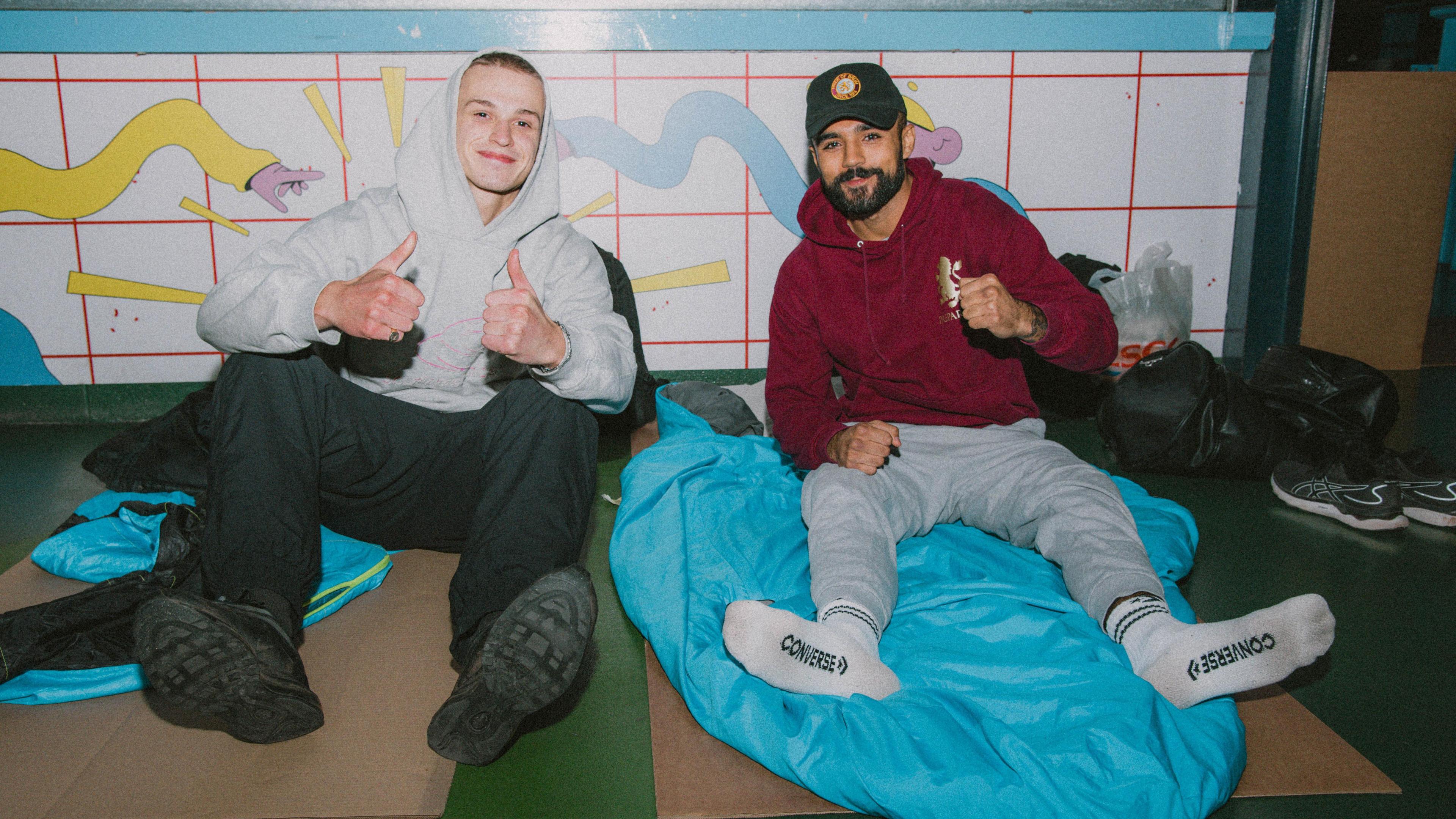 Two men are sitting on the floor with sleeping bags at a previous sleep out. They are making thumbs up signs and are looking happy.