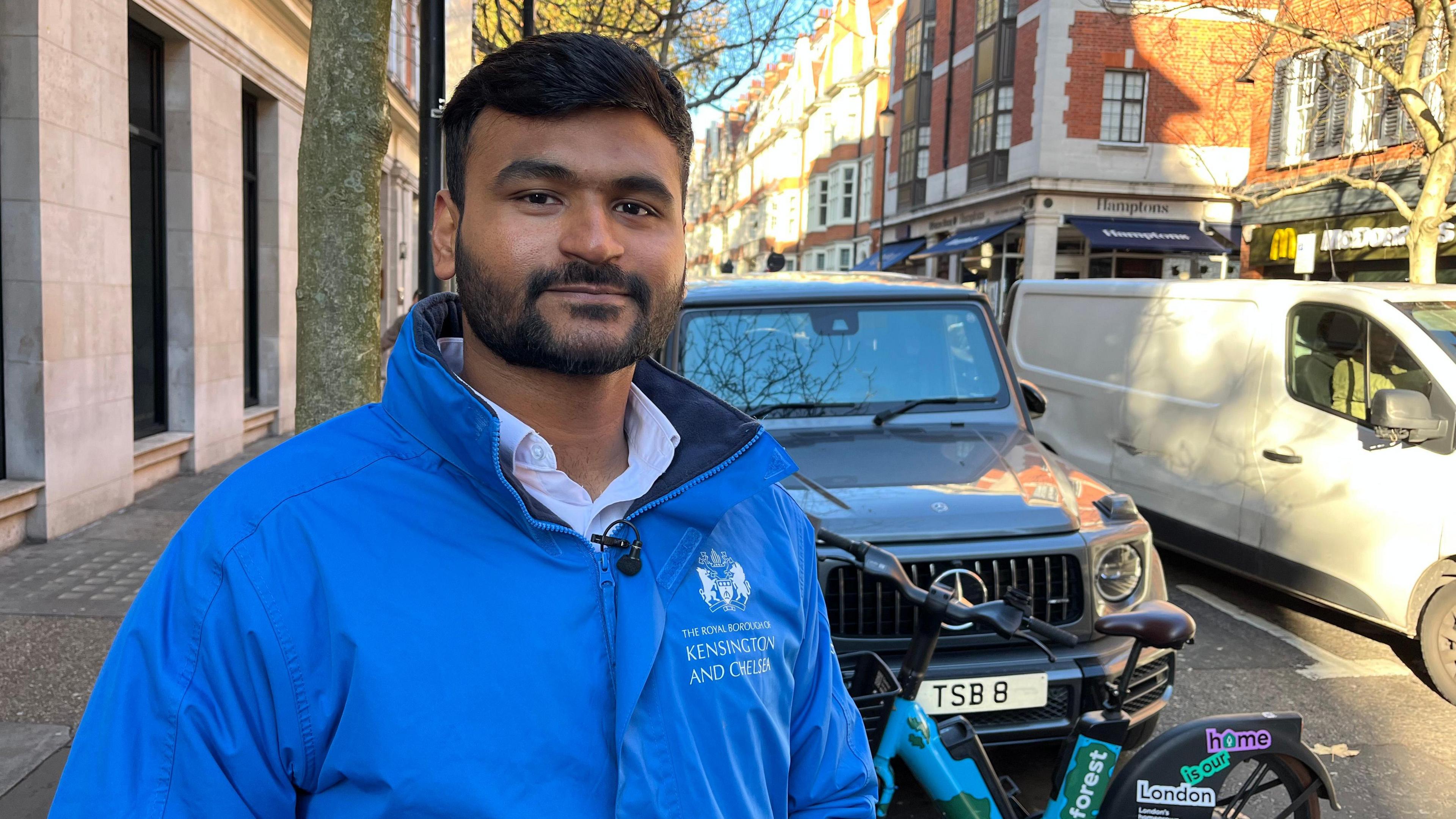 A man in a blue jacket is looking at the camera. In the background a hire bike is parked infront of a jeep.