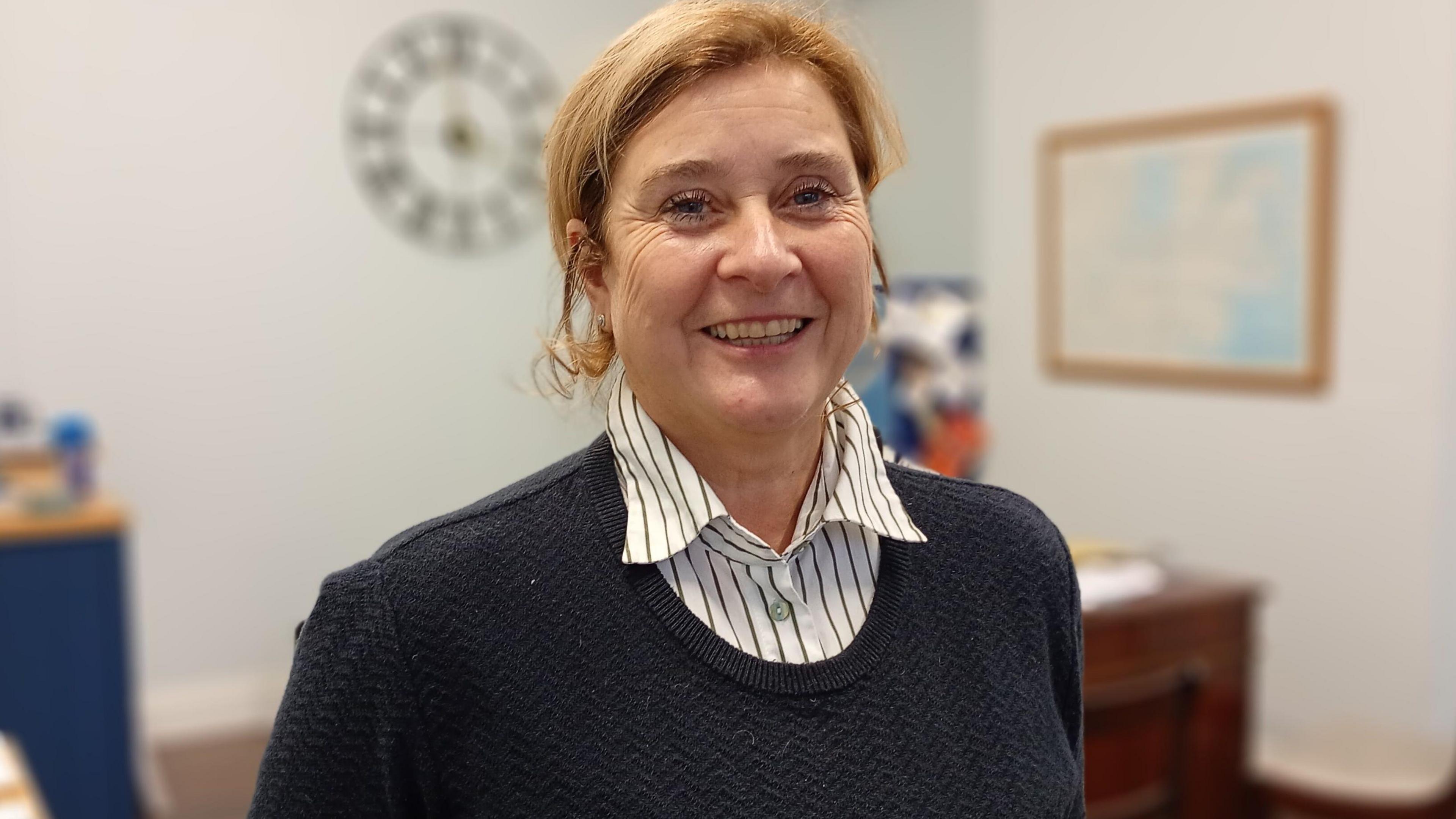 Tanya Graham in a blue jumper and white striped shirt. She is smiling at the camera. She has red hair.