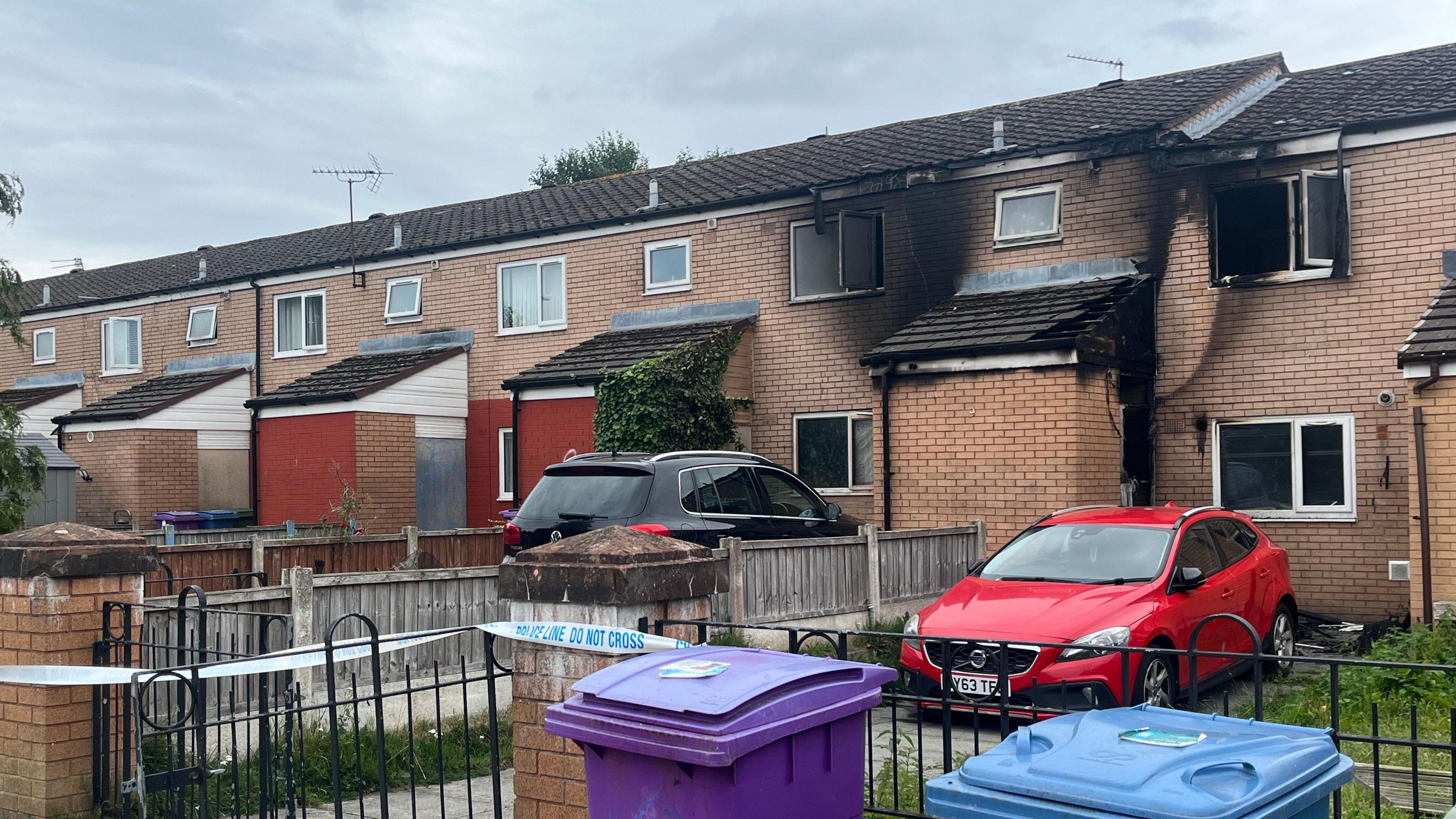 Two terraced houses with visible smoke damage on the brickwork and windows. Both are cordoned off with blue and white police tape. 