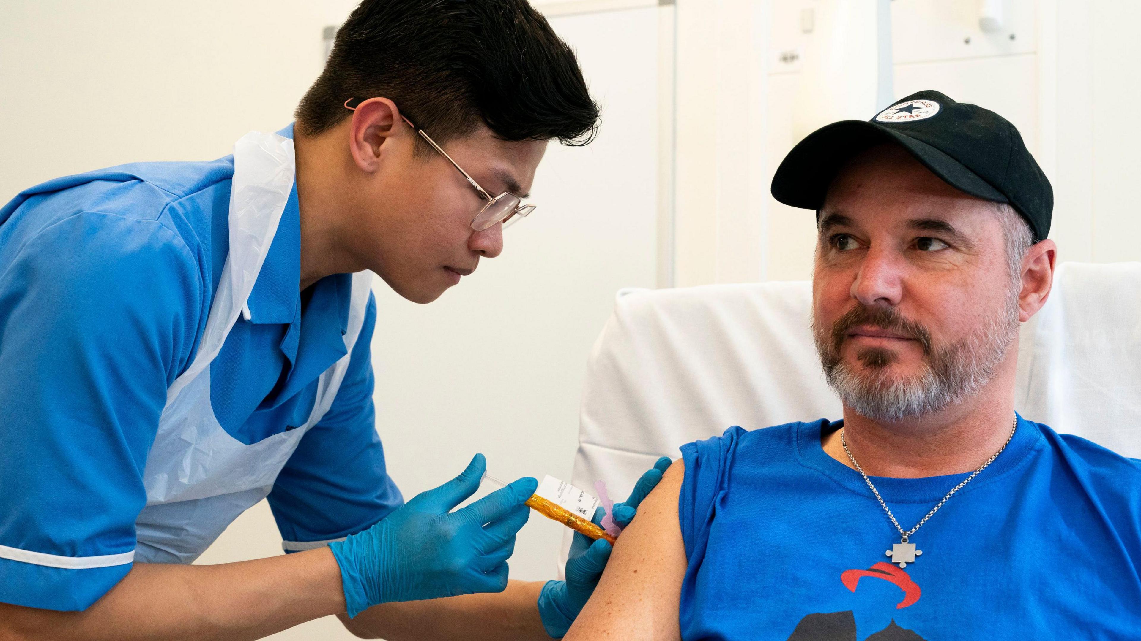 A male nurse wearing a blue uniform and white plastic apron is putting an injection into the arm of Steve Young, who is wearing a blue T-shirt and black baseball cap.