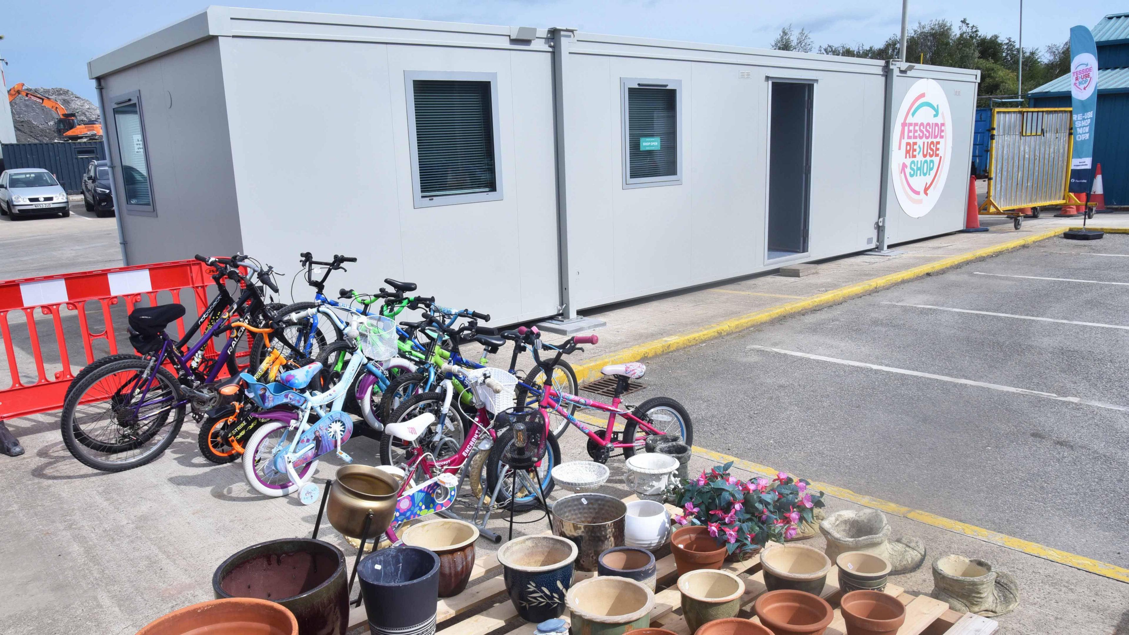 The Teesside Reuse Shop. The shop is located in a grey portable cabin building at the recycling centre.  A selection of children's bicycles stands outside in front of the cabin. in the foreground a selection of plant pots are displayed on a wooden table.