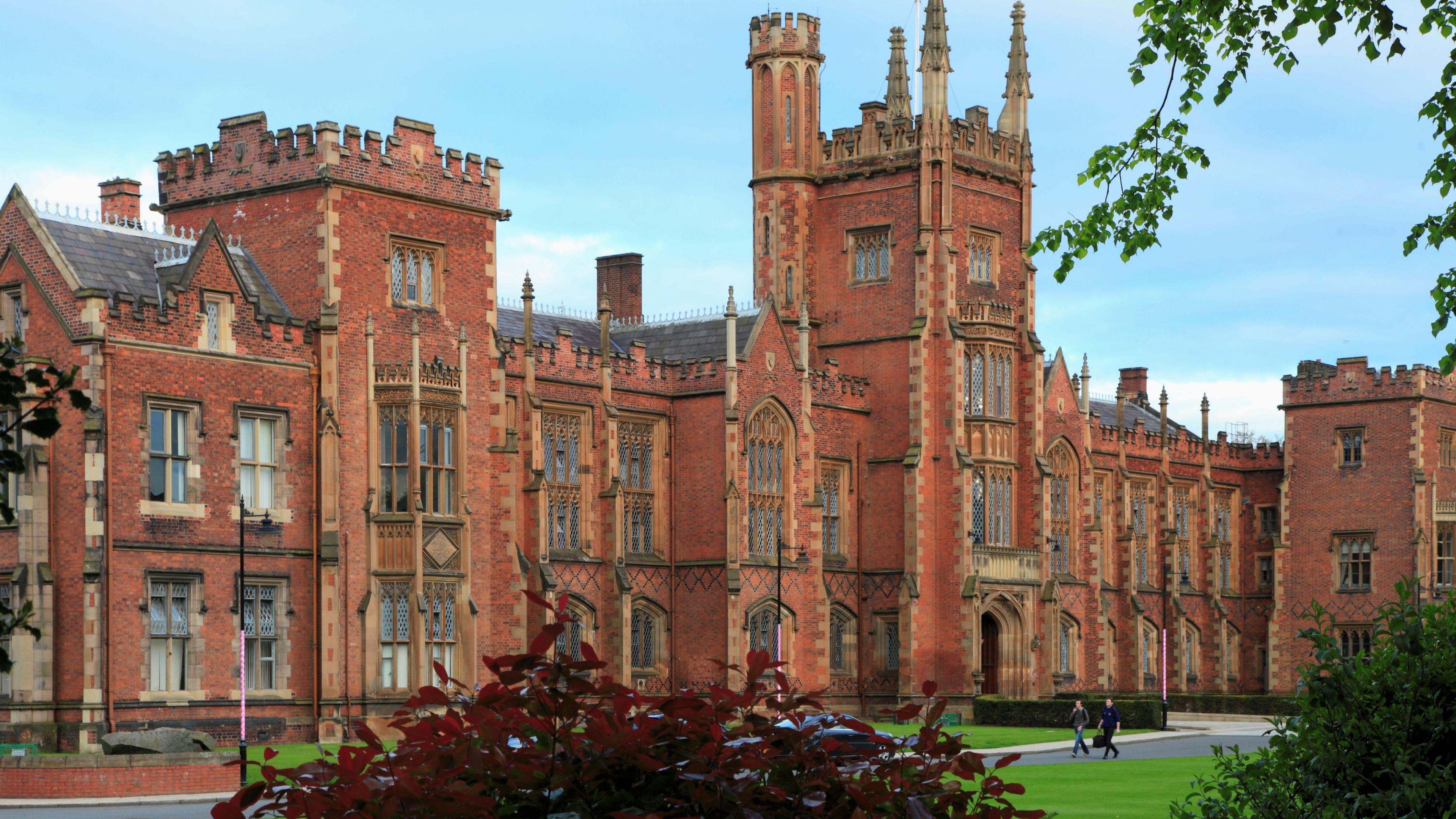 A grand building with red Victorian brick and towers. Two students are walking towards grass and bushes are planted around the building.