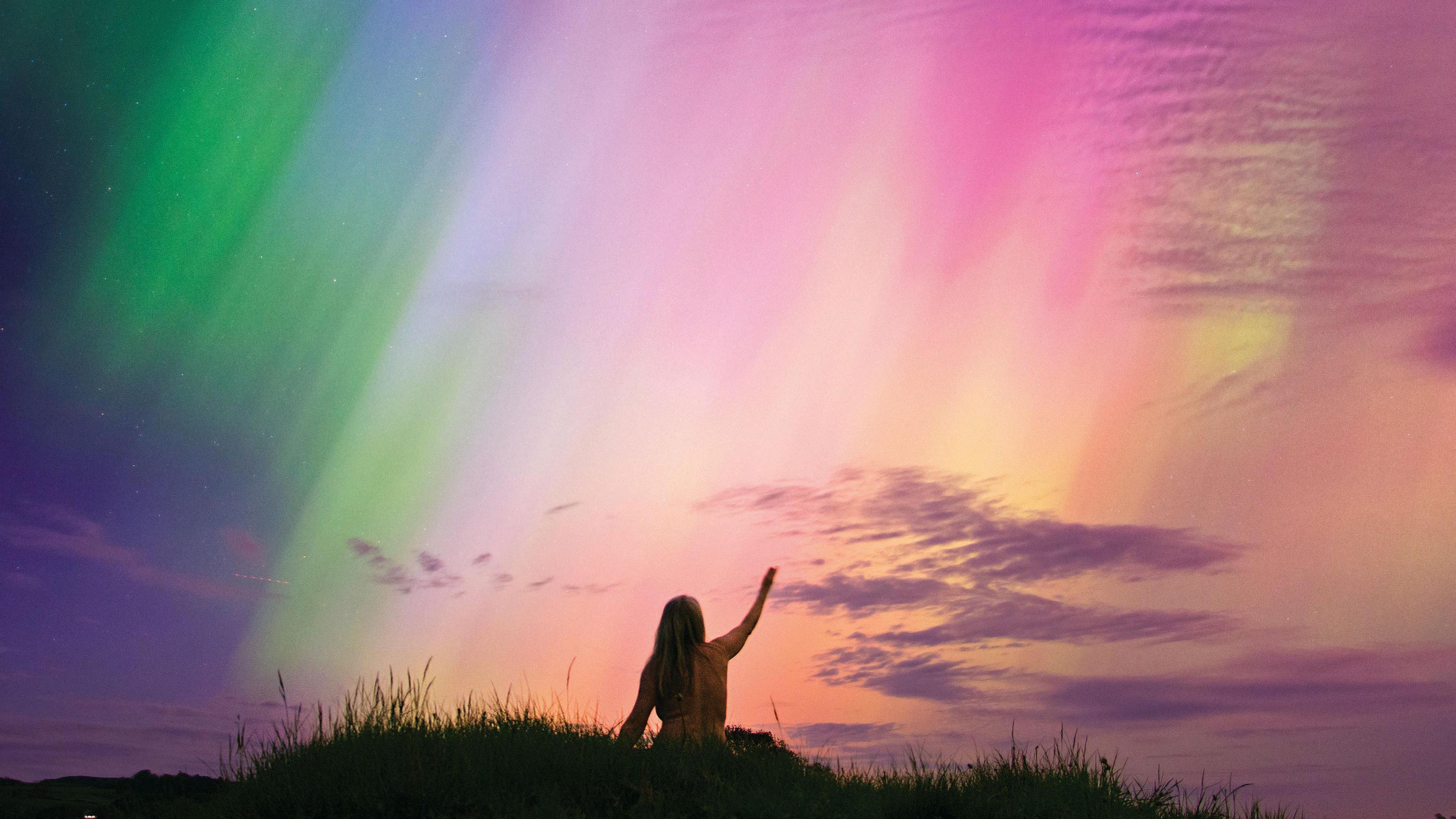 The image shows the back of a naked woman with her arm raised to the sky against a backdrop of the northern lights 