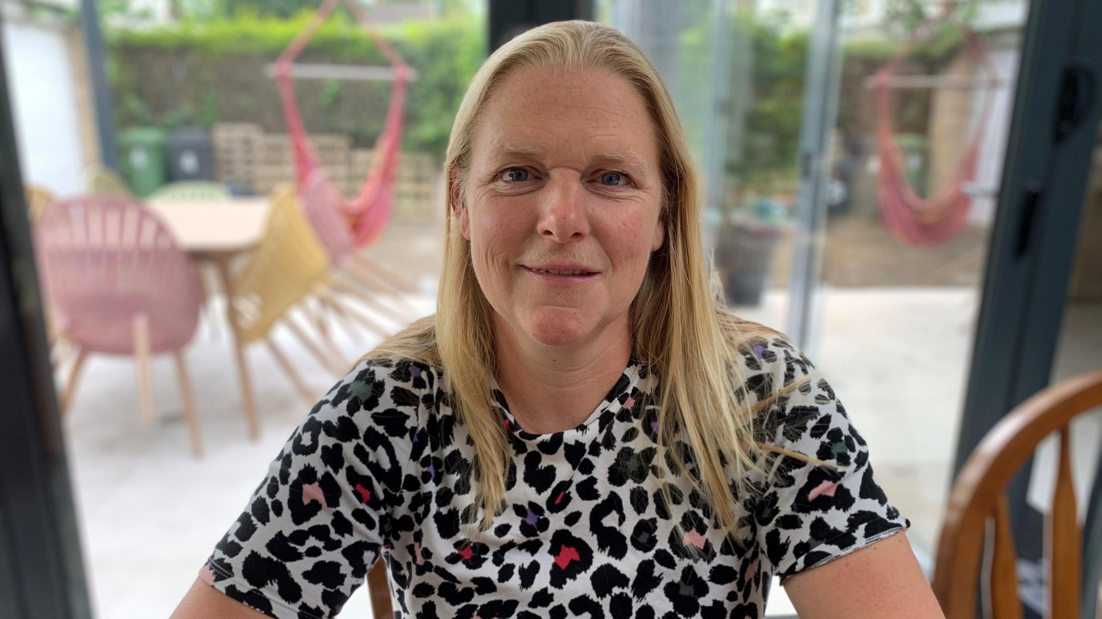 A woman in her early 50s with shoulder length blond hair smiling and wearing a colourful animal print t-shirt sat at a kitchen table.
