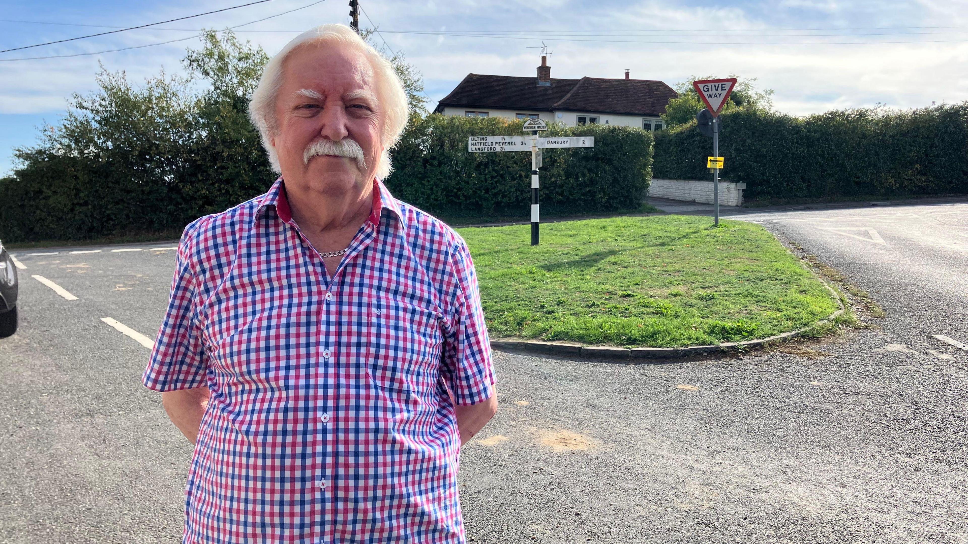 Peter Gaywood looks directly at the camera with a serious expression. He is wearing a red and blue plaid shirt and a silver chain. He is photographed with his hands behind his back standing on a road with a small median strip behind him. 