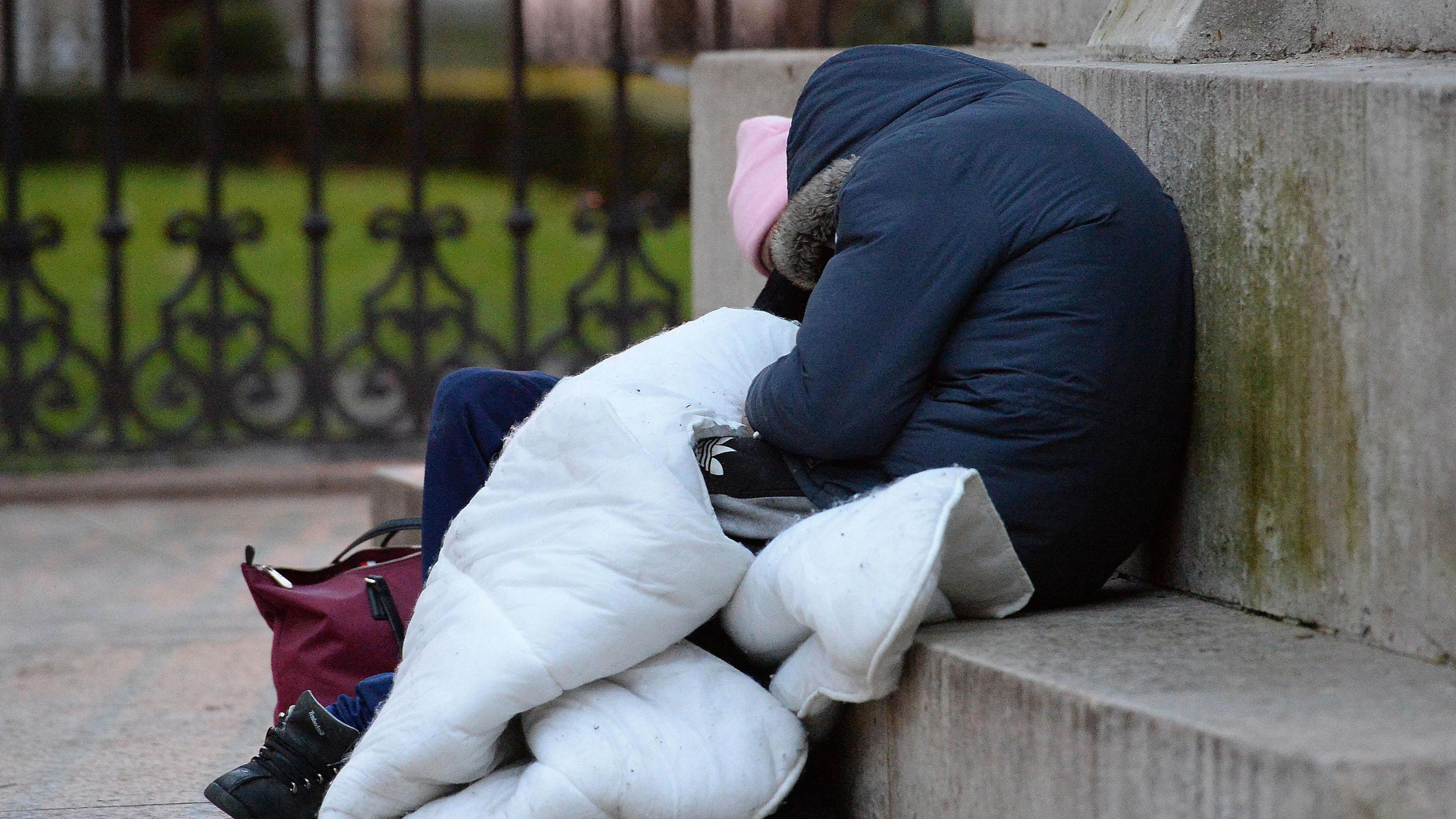 Two people sat on a concrete seat. Both are hunched over in thick jackets with their hoods up. Their legs are covered with a duvet to keep them warm. 