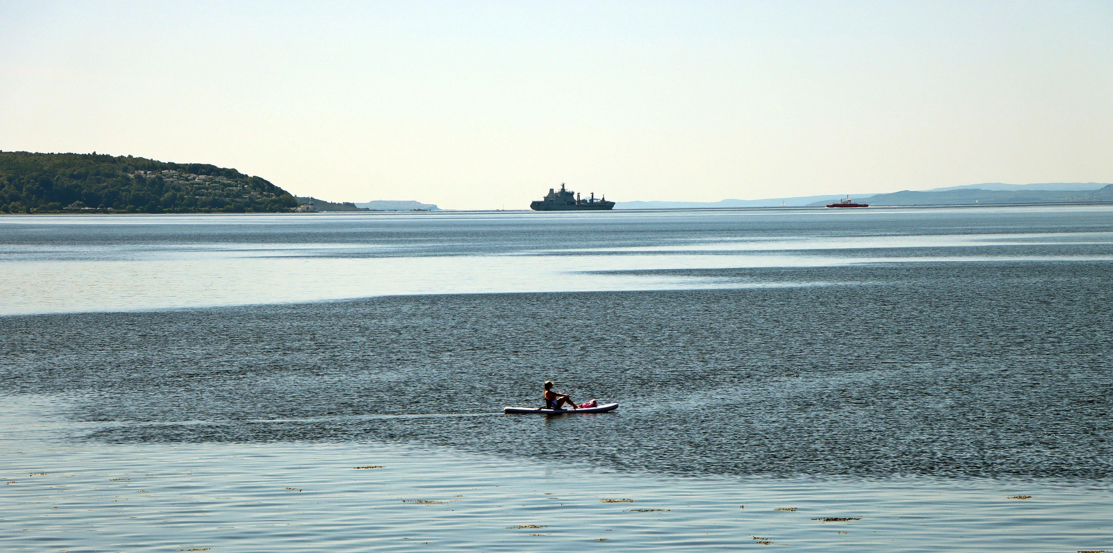 A person in a canoe in the foreground with a large navy ship in the far distance