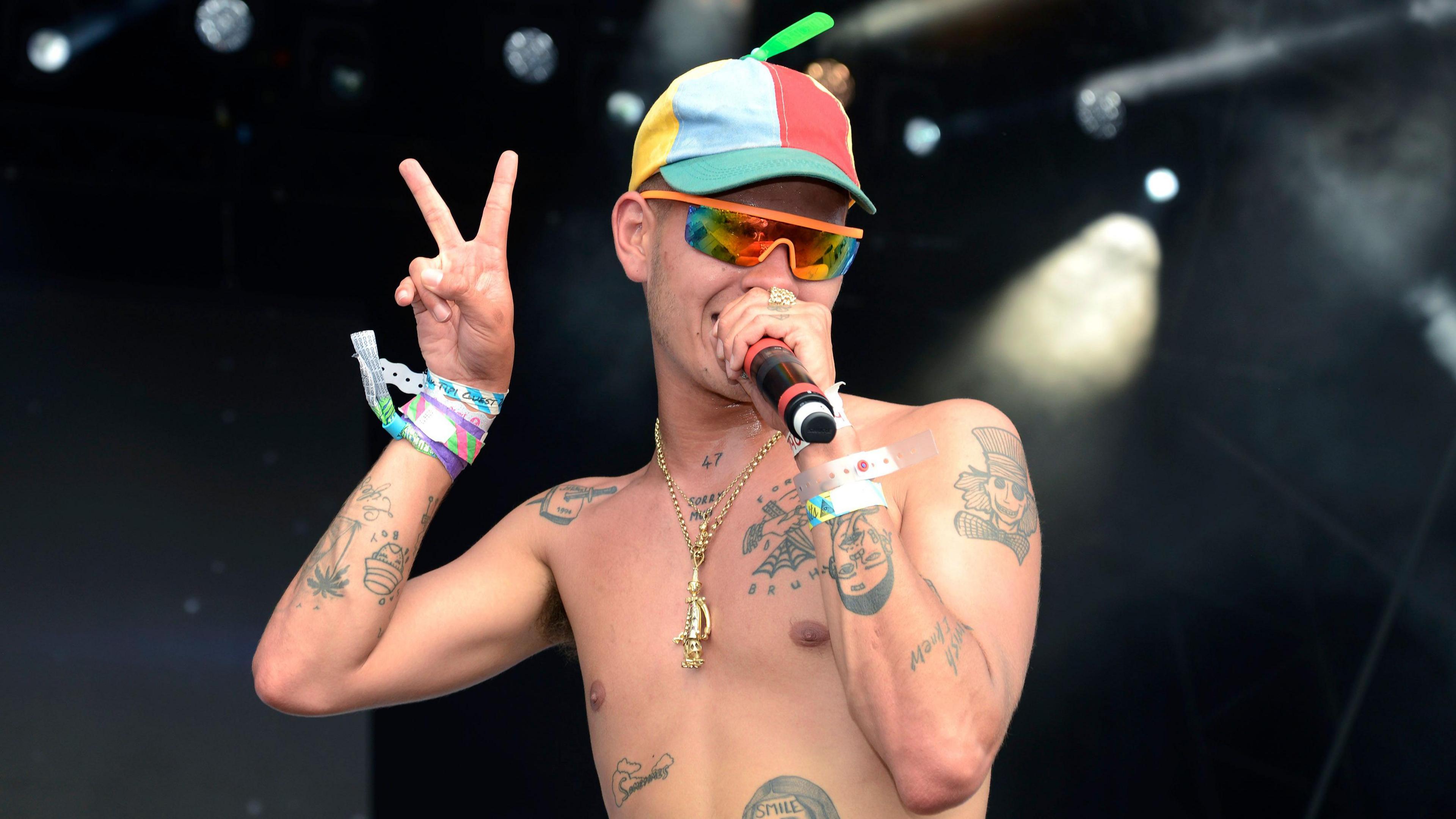 Slowthai performing at a festival. He is wearing a multicoloured hat and sunglasses and is singing into a microphone. He is shirtless, showing several tattoos on his torso and arms. He is making a peace sign with his right hand.