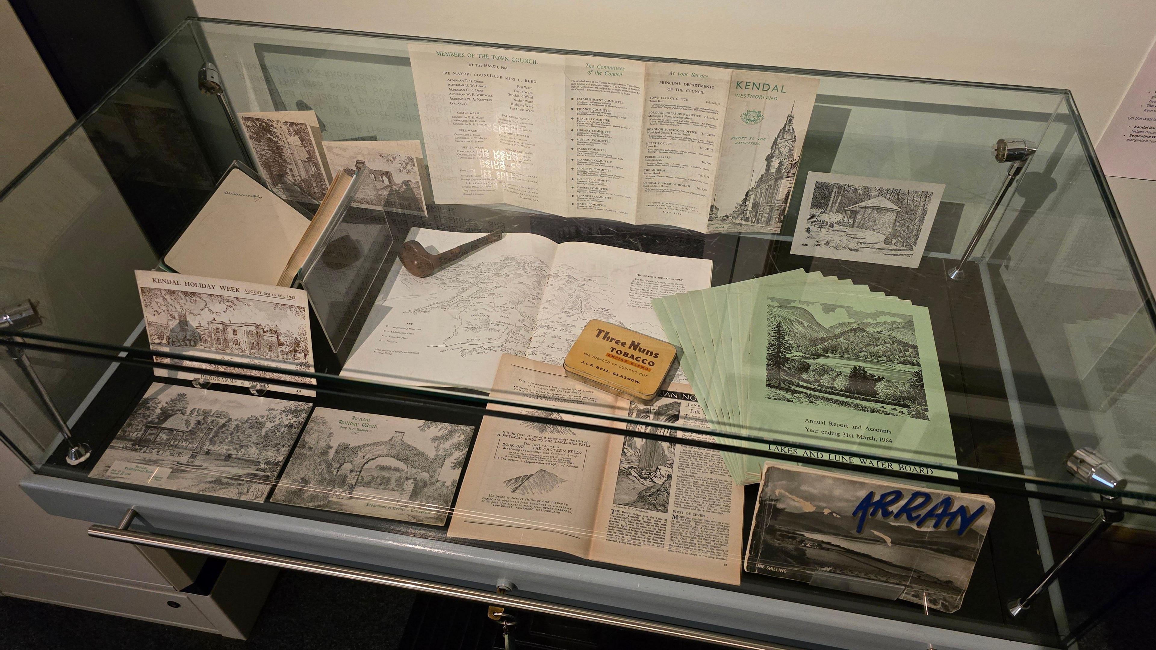 A glass display case containing pen-lined drawings, a wooden smoking pipe and a tobacco tin.