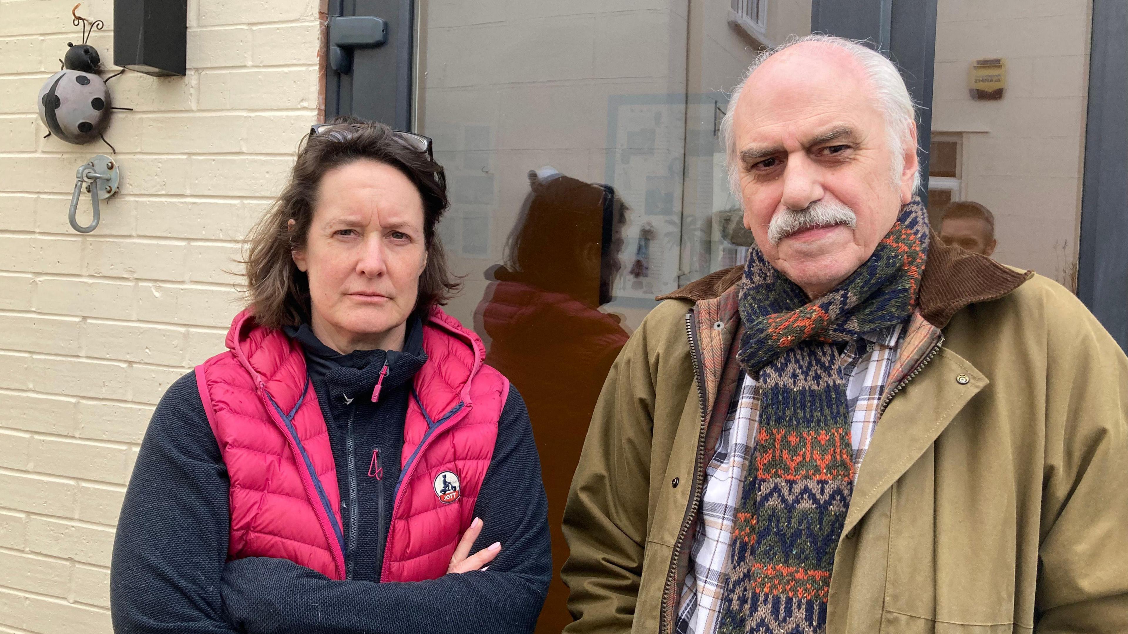 Emma Chetwynd Stapylton standing next to Nick Morrow-Brown outside of her garden office. Emma has medium-length brown hair with glasses resting on top of her head. She wears a pink gilet over a fleece and has her arms crossed. Nick wears a thick brown jacket, a checked shirt and a wool scarf. He looks at the camera with a stern face.