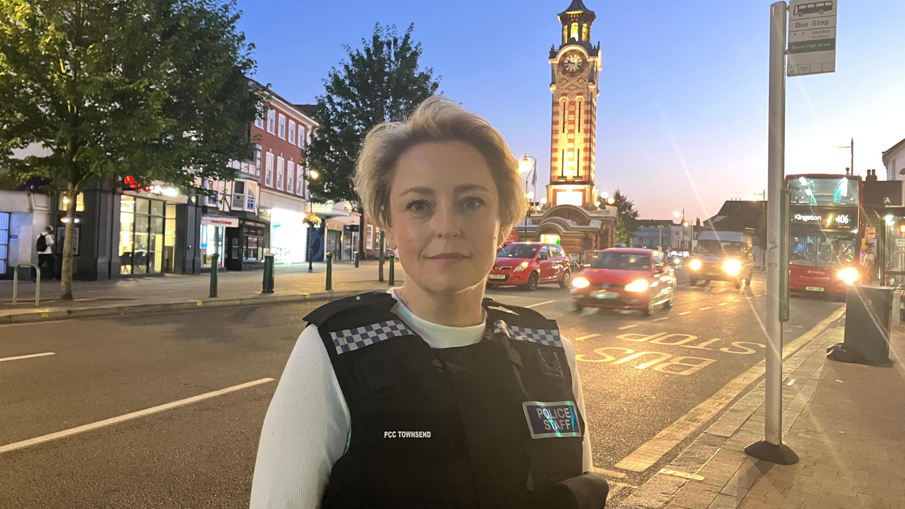 Police and Crime Commissioner Lisa Townsend looks at the camera, she is wearing a police vest with a police staff label on. In the background there is a clock tower which is lit up in the evening light and cars driving past with their headlights on.