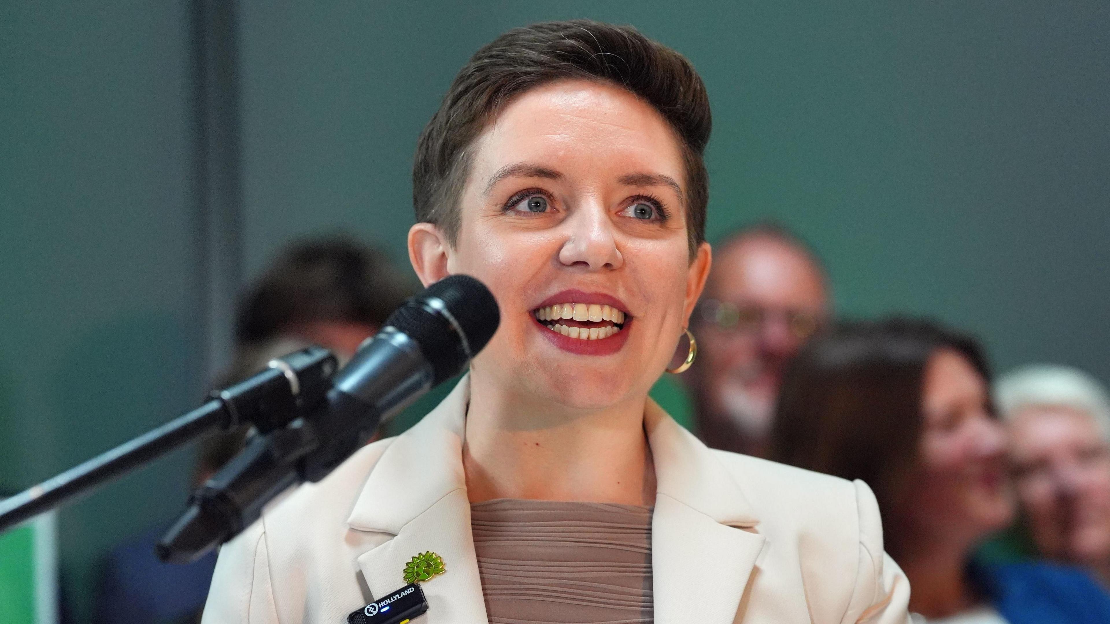 Carla Denyer standing behind a microphone ready to speak. She is wearing a cream jacket and brown top, with red lipstick and gold hoop earrings.