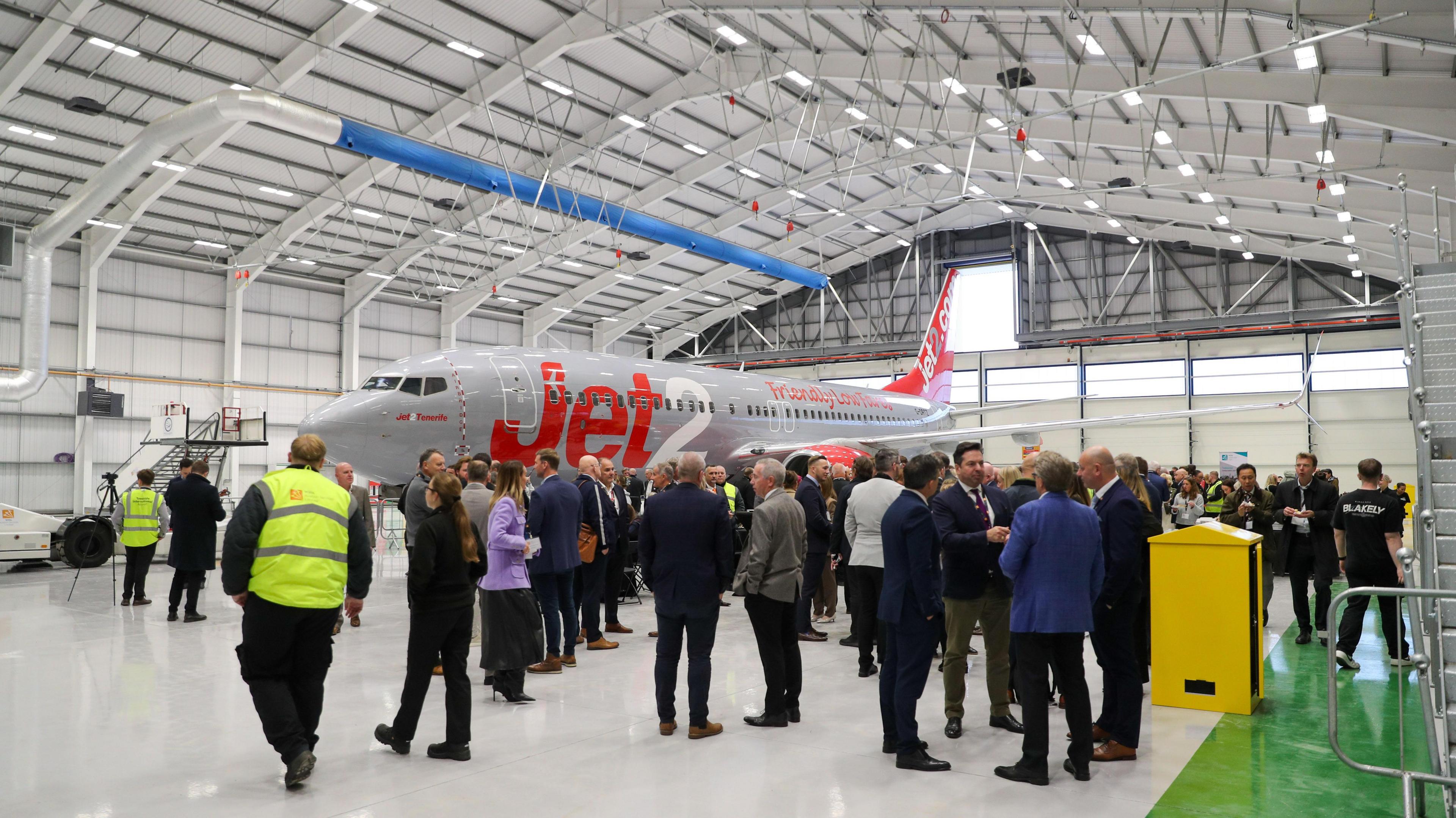 Dozens of guests standing in the new hanger. They are gathered next to a Jet2 plane, which has a grey and red livery and the firm's logo on the side.