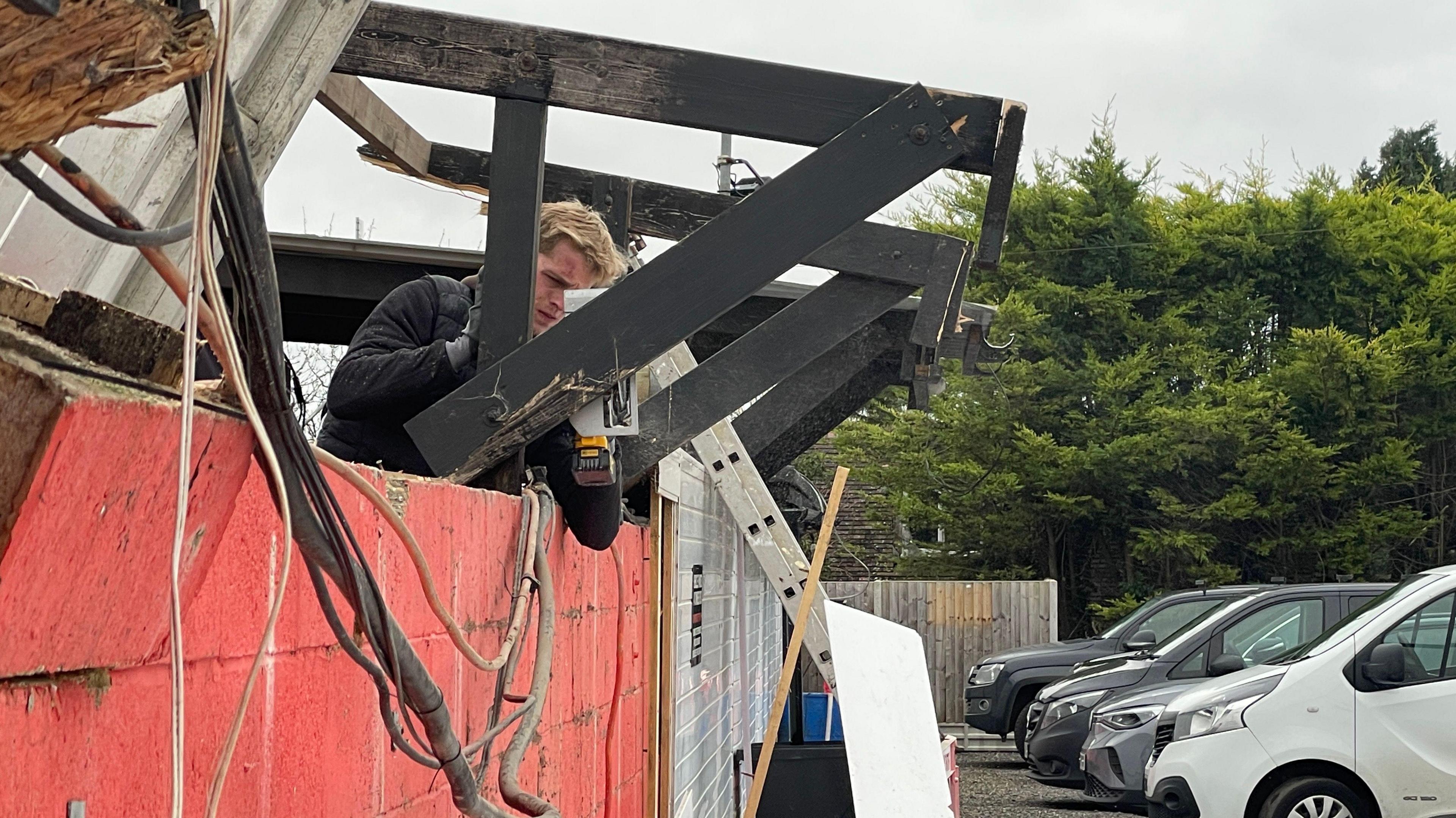 A broken roof with a man trying to fix it. There are cars parked on the right of the picture. There is a ladder leaning against the building and the roof has been ripped off.