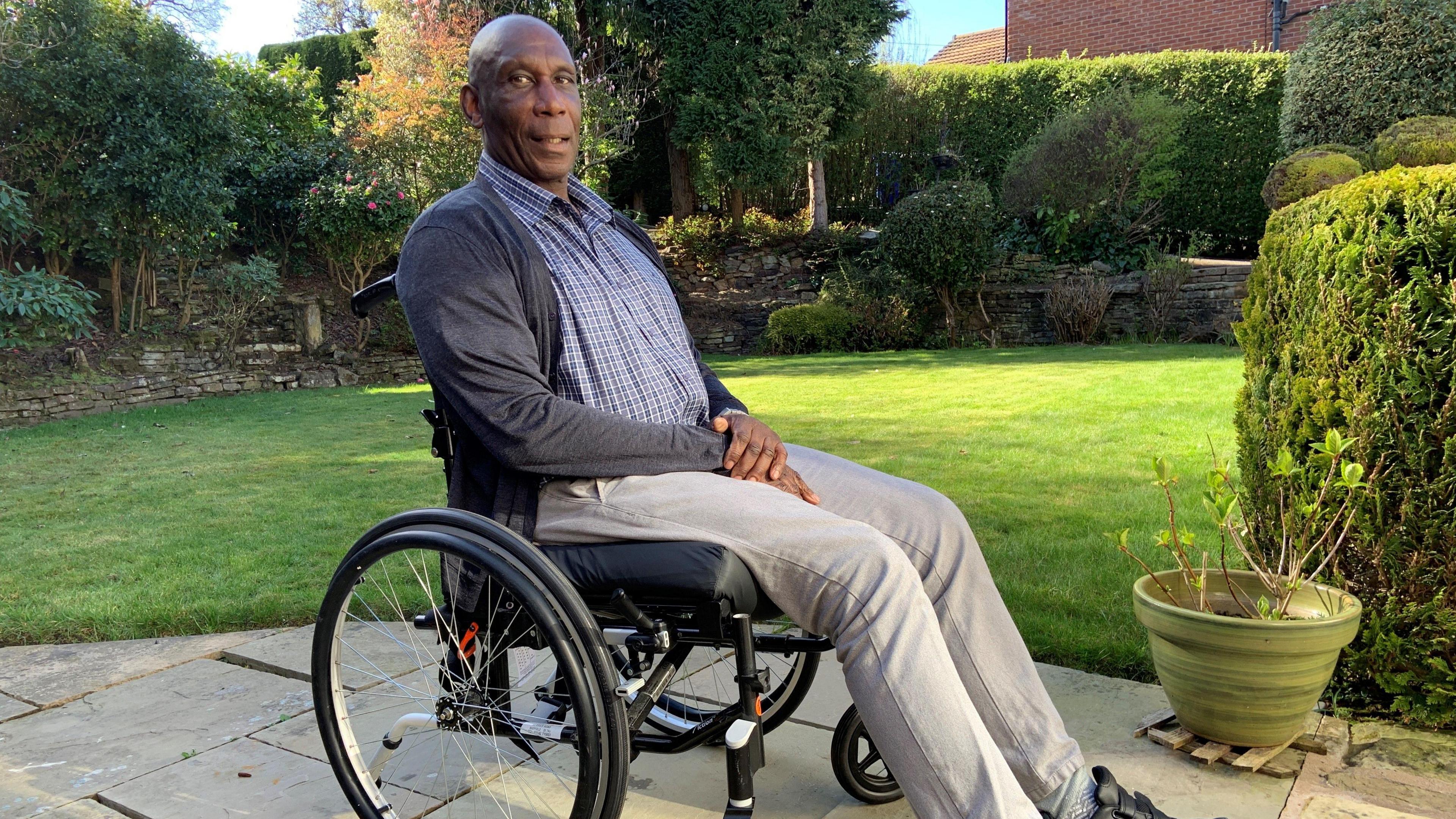 Uriah Rennie sits in a wheelchair. He is sitting sideways and looking into the camera. He is wearing a shirt, cardigan and trousers
