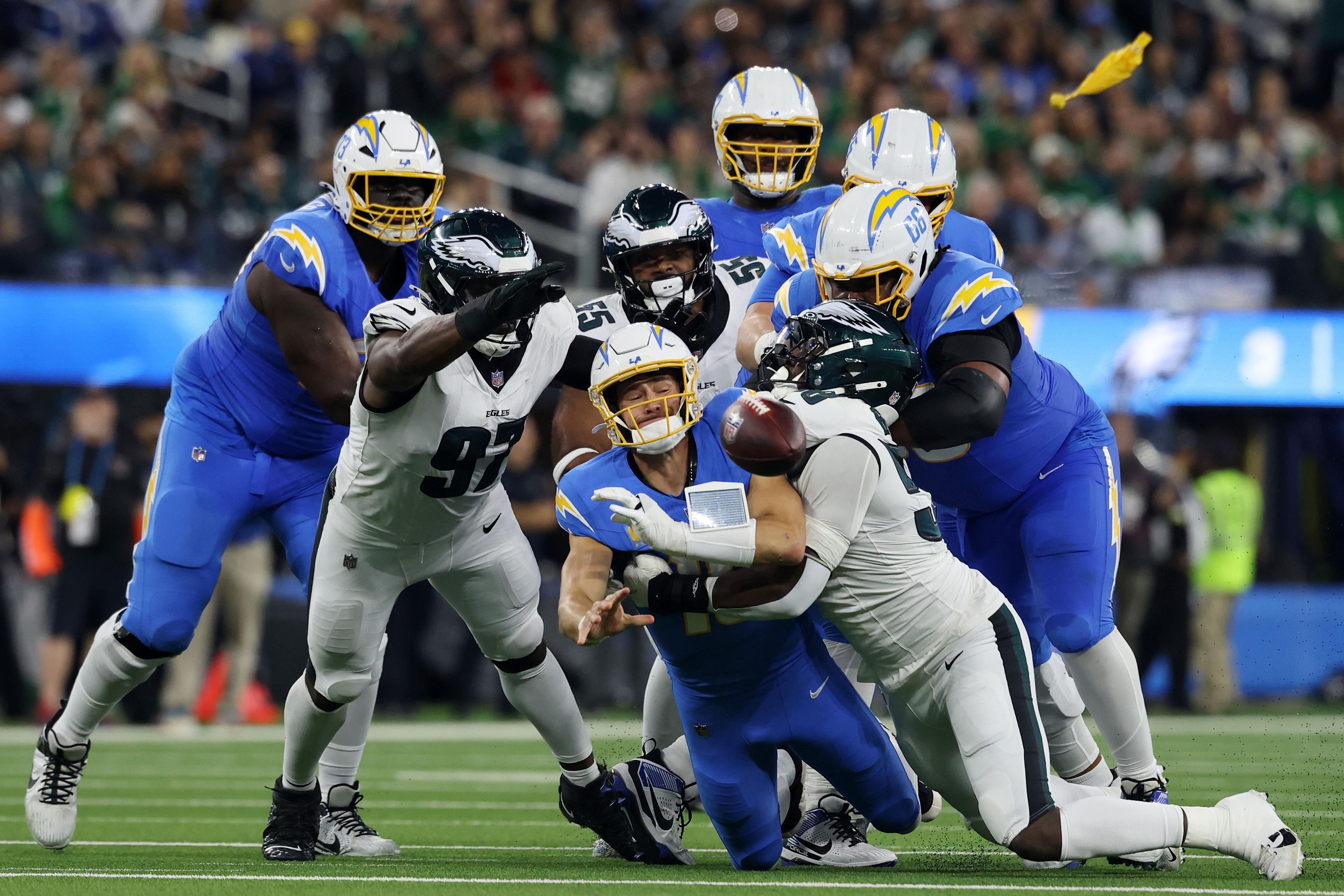 Players from the Philadelphia Eagles tackle the Los Angeles Chargers quarterback as the ball pops loose, surrounded by several Chargers linemen attempting to protect the play during an intense moment on the field.
