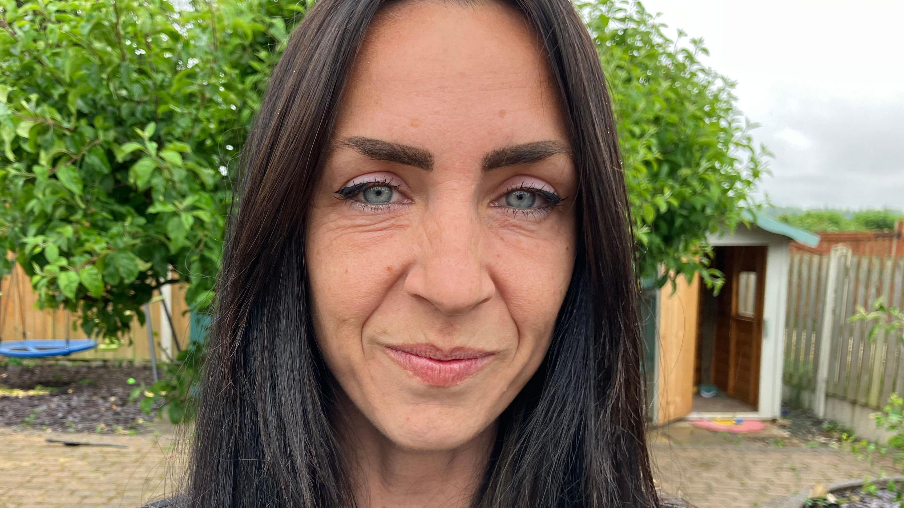 A woman with long brown hair stands in a garden. She is smiling at the camera.