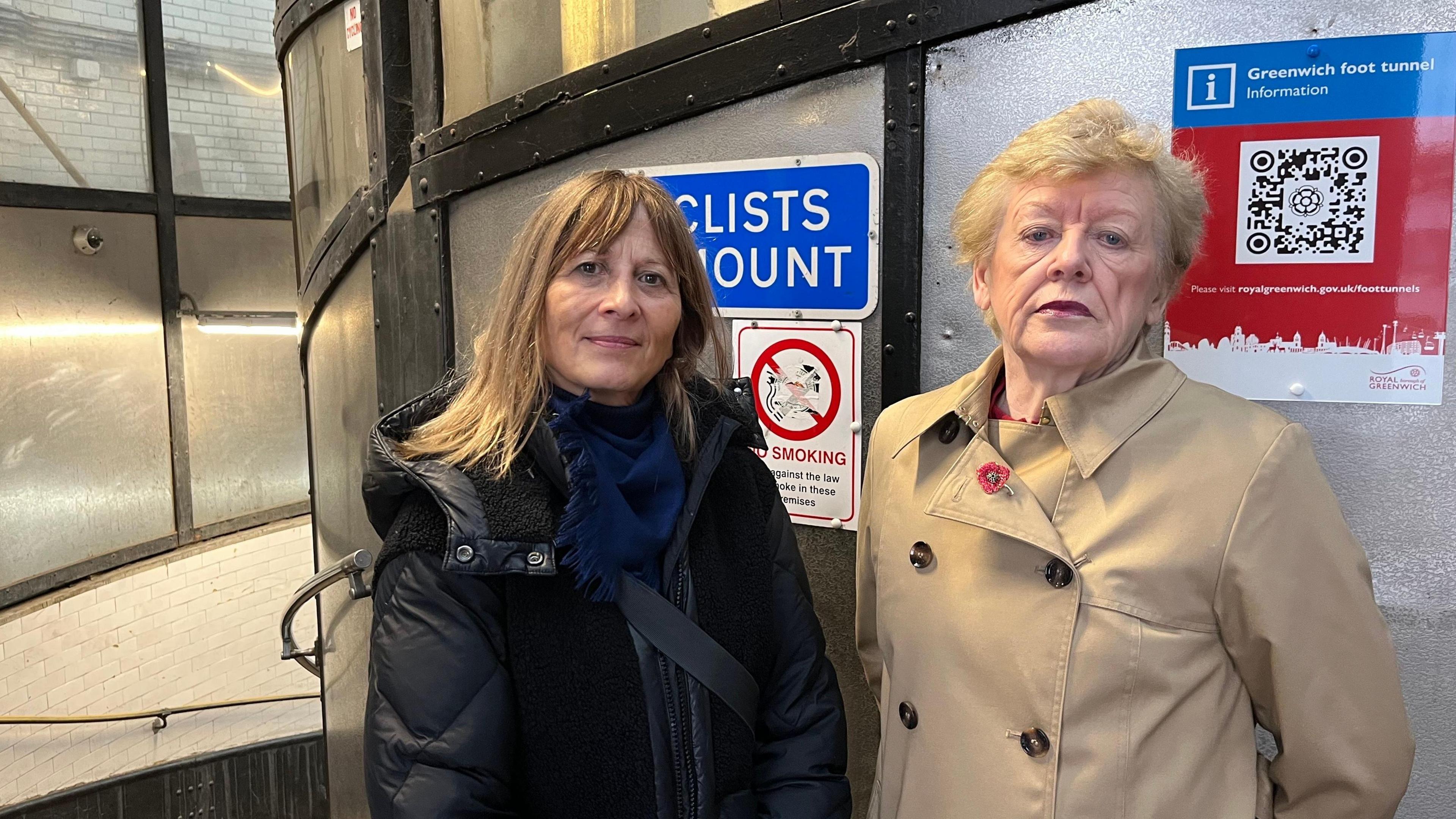 Two women stood next to each other in front of a metal wall, at the top of a stairwell. They both have a straight face and are wearing jackets.