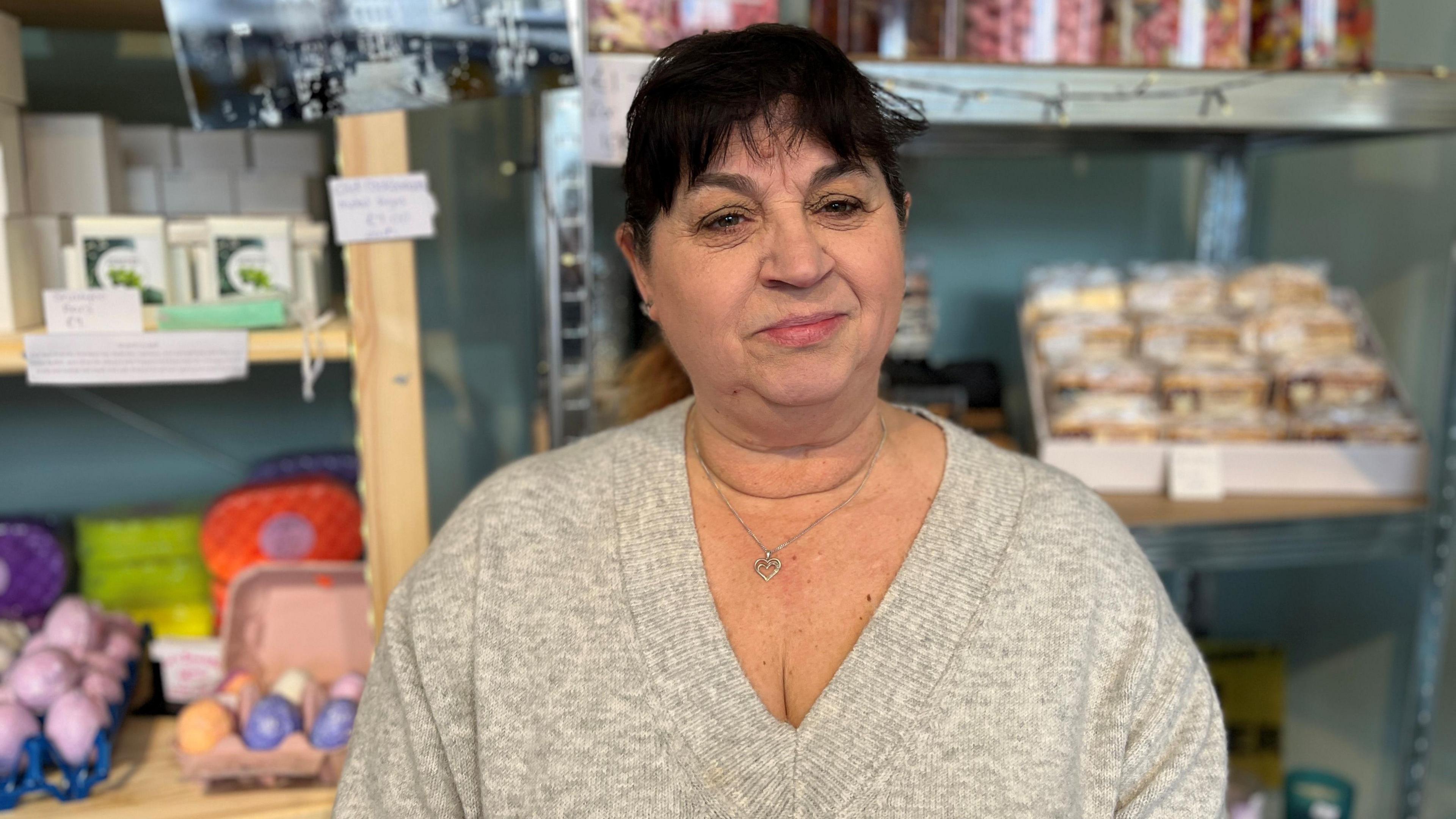 Lorraine Barrett standing in front of a display in her gift shop in Cockermouth. She has dark hair in a pony tail with a fringe. She is wearing a grey jumper and a silver necklace with a heart pendant.