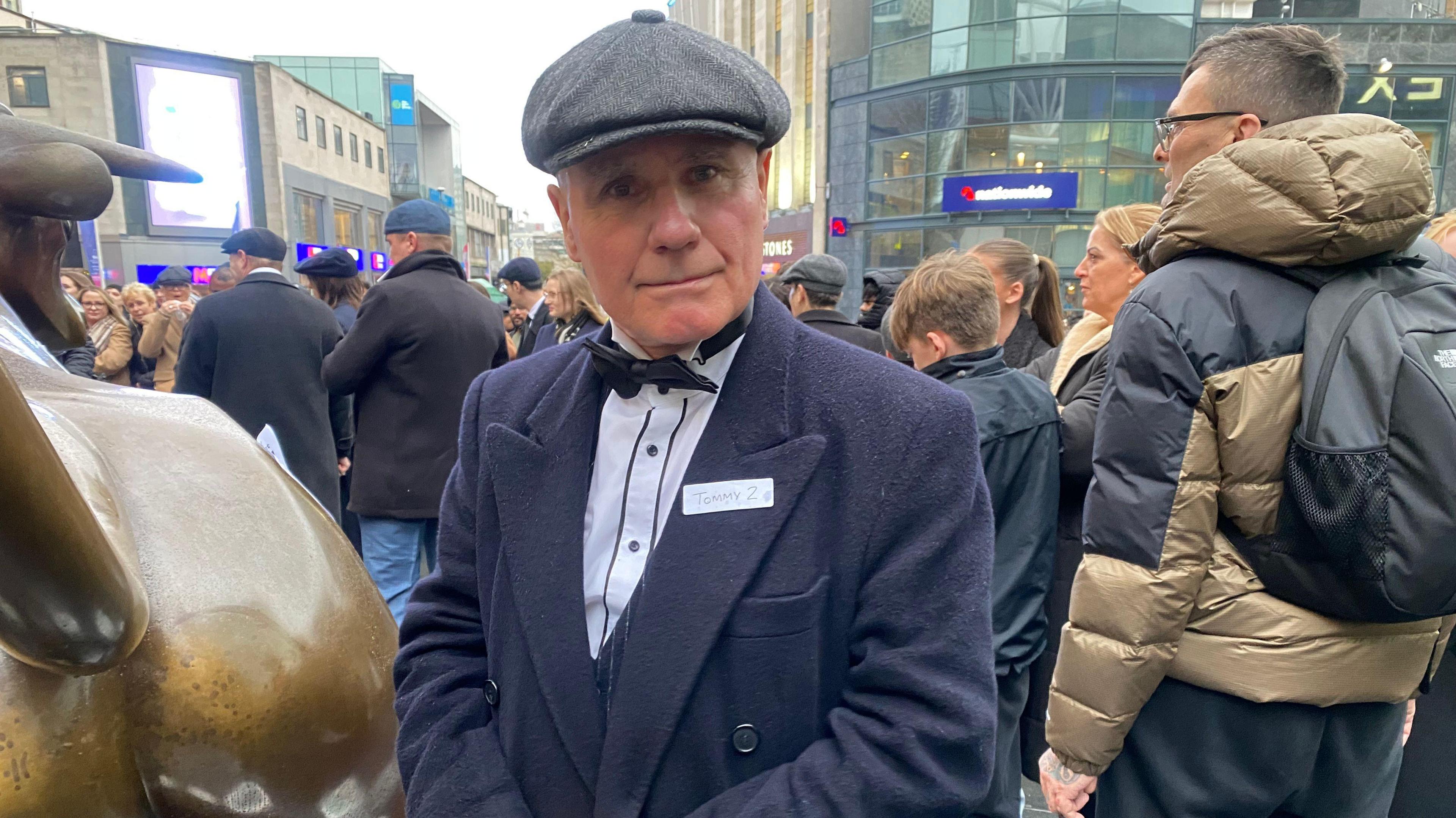 a man wearing a black flat cap and black overcoat with a black bow tie on a white shirt