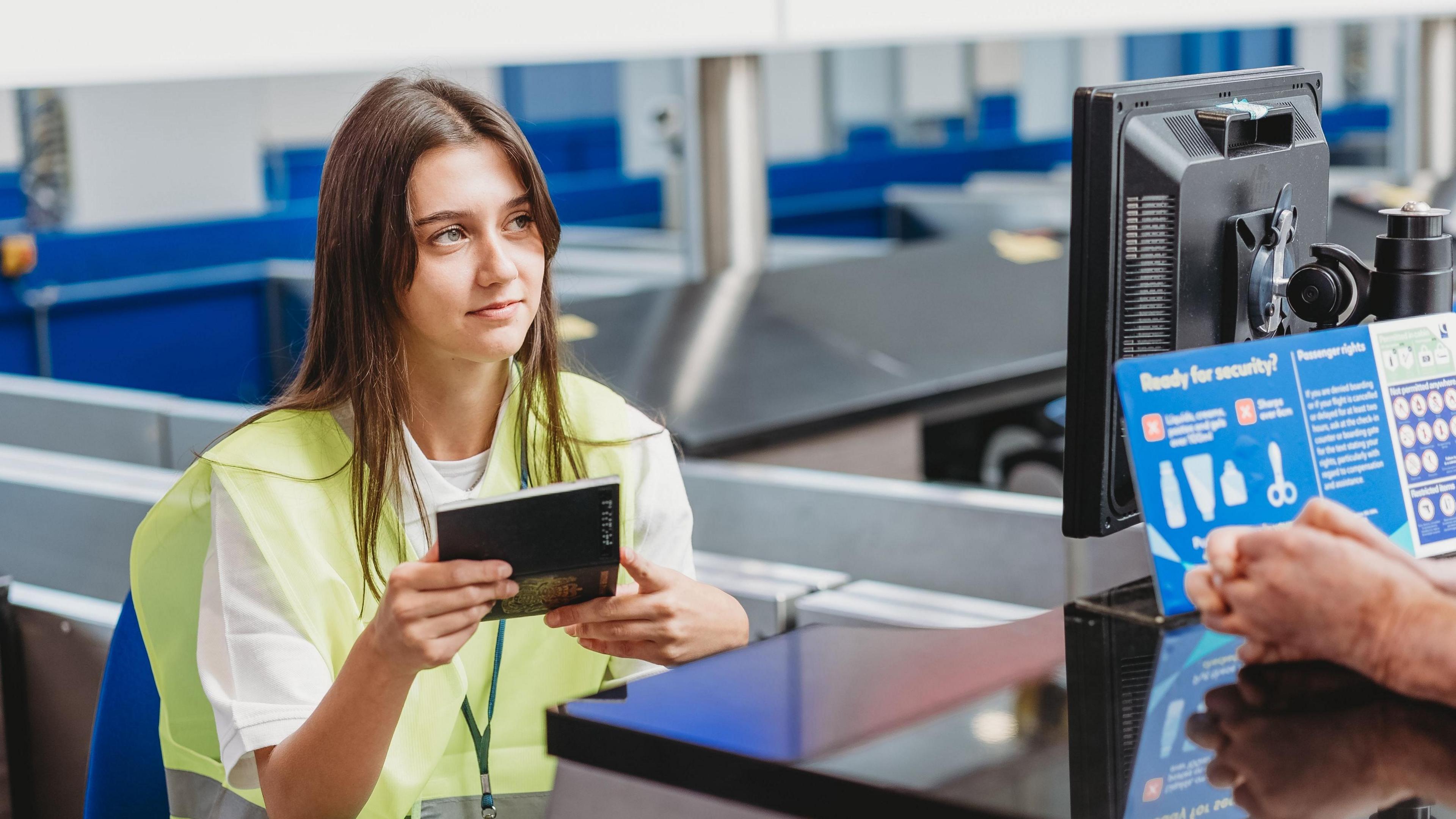 A young woman holding a passport wearing a high-vis jacket at an airport. She is looking towards another individual who has their hands on the table of the check-in point. There is a blue poster in the foreground as well as a computer.