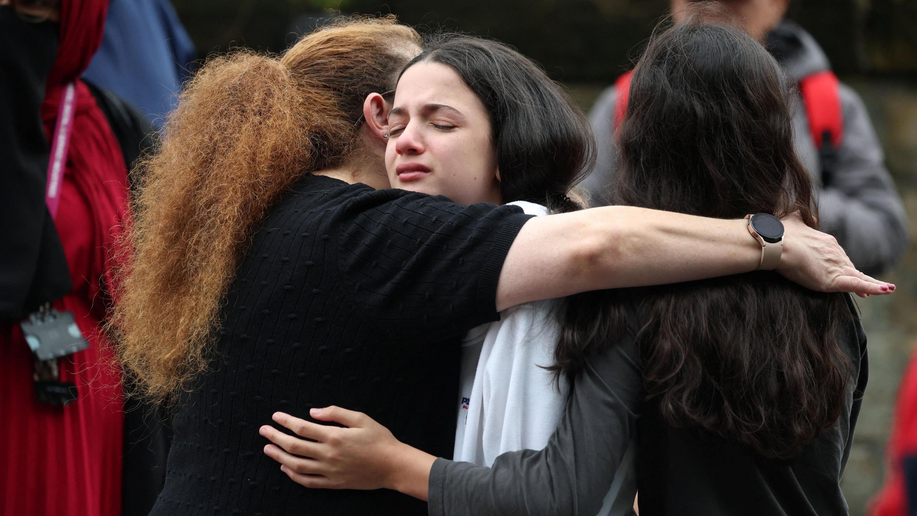 A young girl with a pained face hugs two other women. She has long dark hair and wears white. The woman on her left wearing black has red, curly hair and the woman to her right wearing grey has dark, wavy hair. Several others stand around them.
